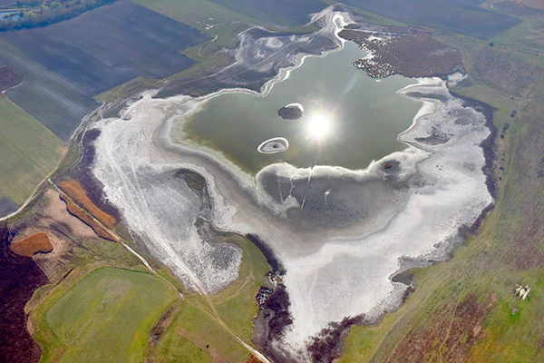 Aerial view of a salt lake with white salt deposits surrounding a central green body of water, surrounded by agricultural fields and green landscapes