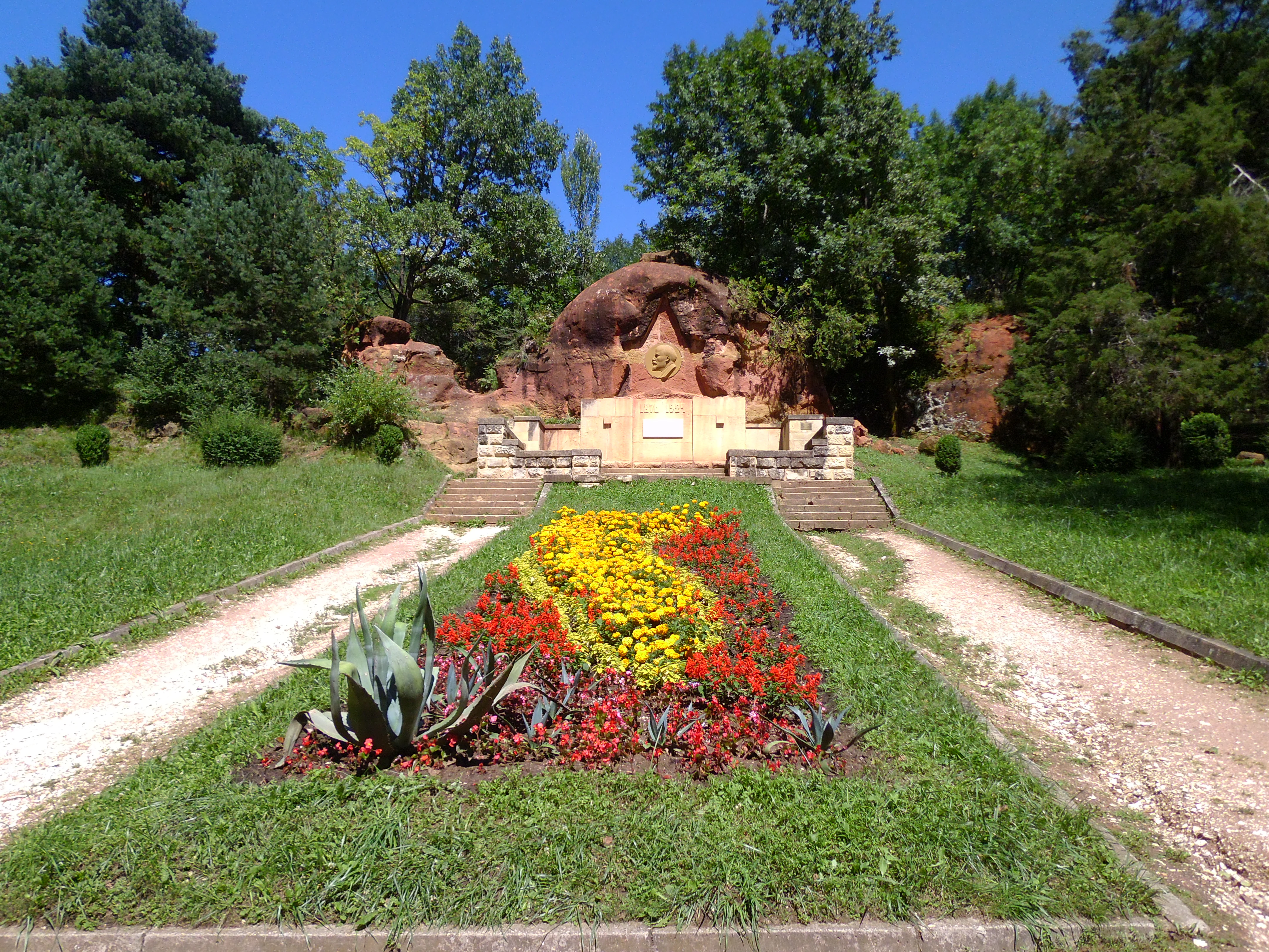 A wide angle shot of the Red Stones rock formation behind a formal flower garden with red, yellow, and green plants, with a walking path on either side and trees in the background.
