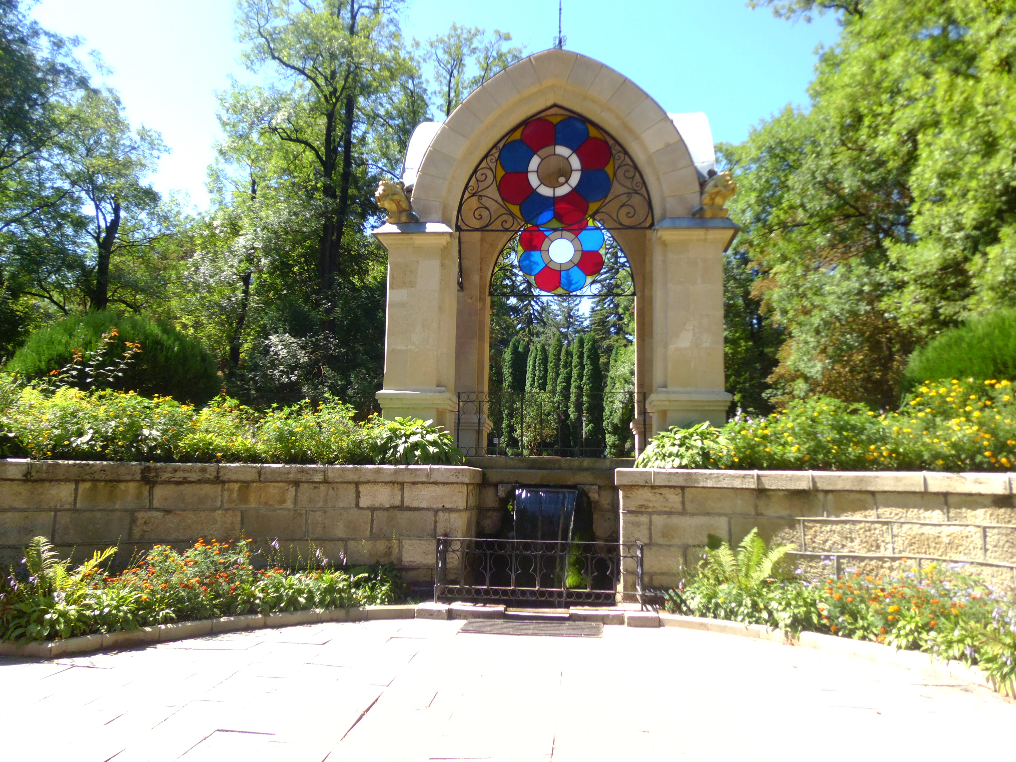 A decorative arched stone pavilion with a stained glass floral insert, situated above a small water feature in a garden setting.
