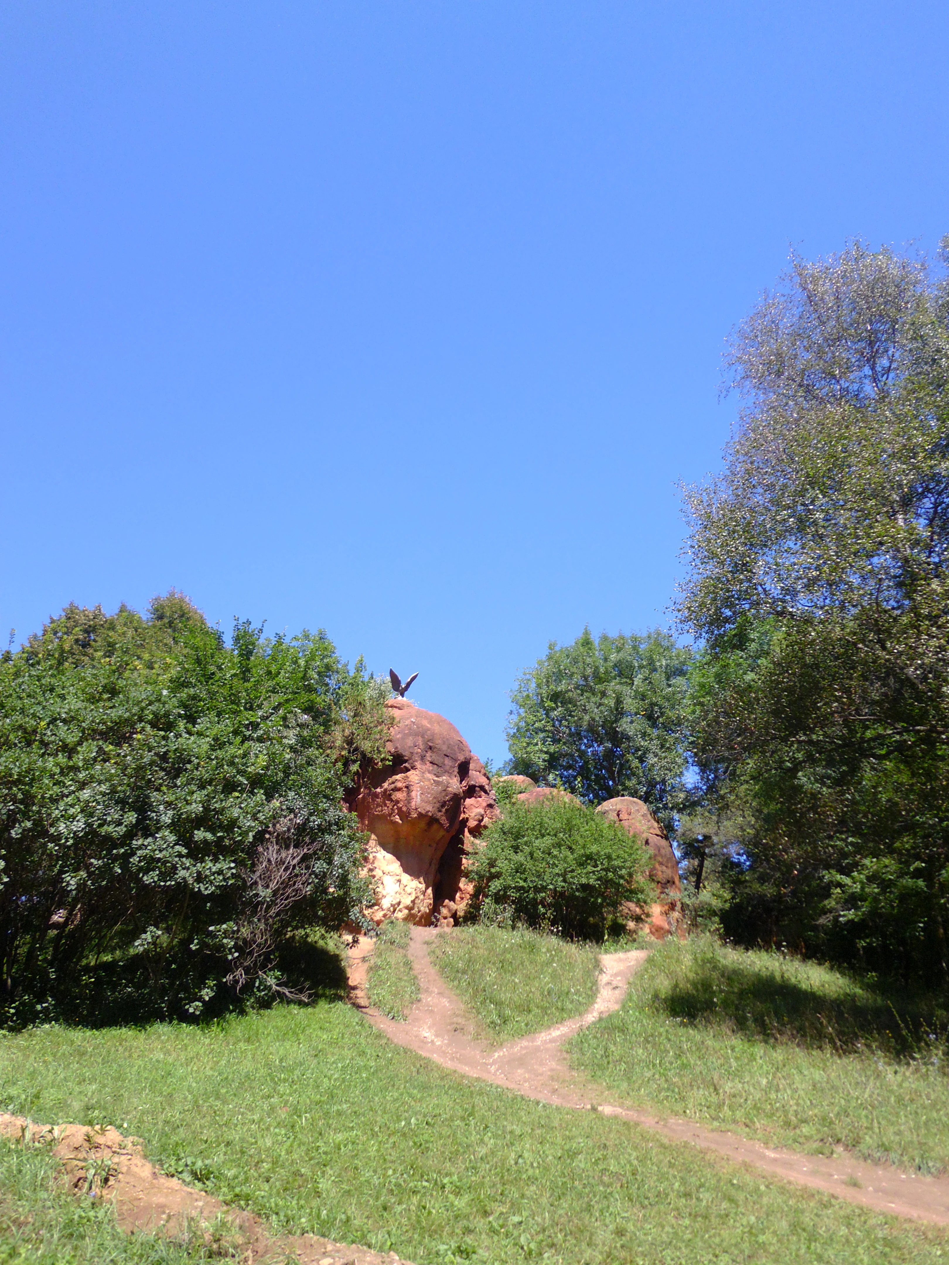 A bronze eagle sculpture perched on top of a reddish sandstone rock formation, surrounded by green trees and grass with a dirt path in the foreground under a clear blue sky.