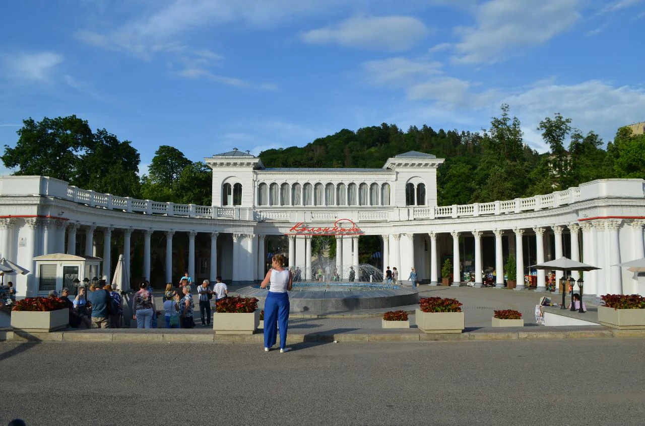 A wide shot of the grand, white, neoclassical-style colonnade at the entrance of Kislovodsk National Park, featuring a semicircular row of columns and people walking in the paved plaza in front.