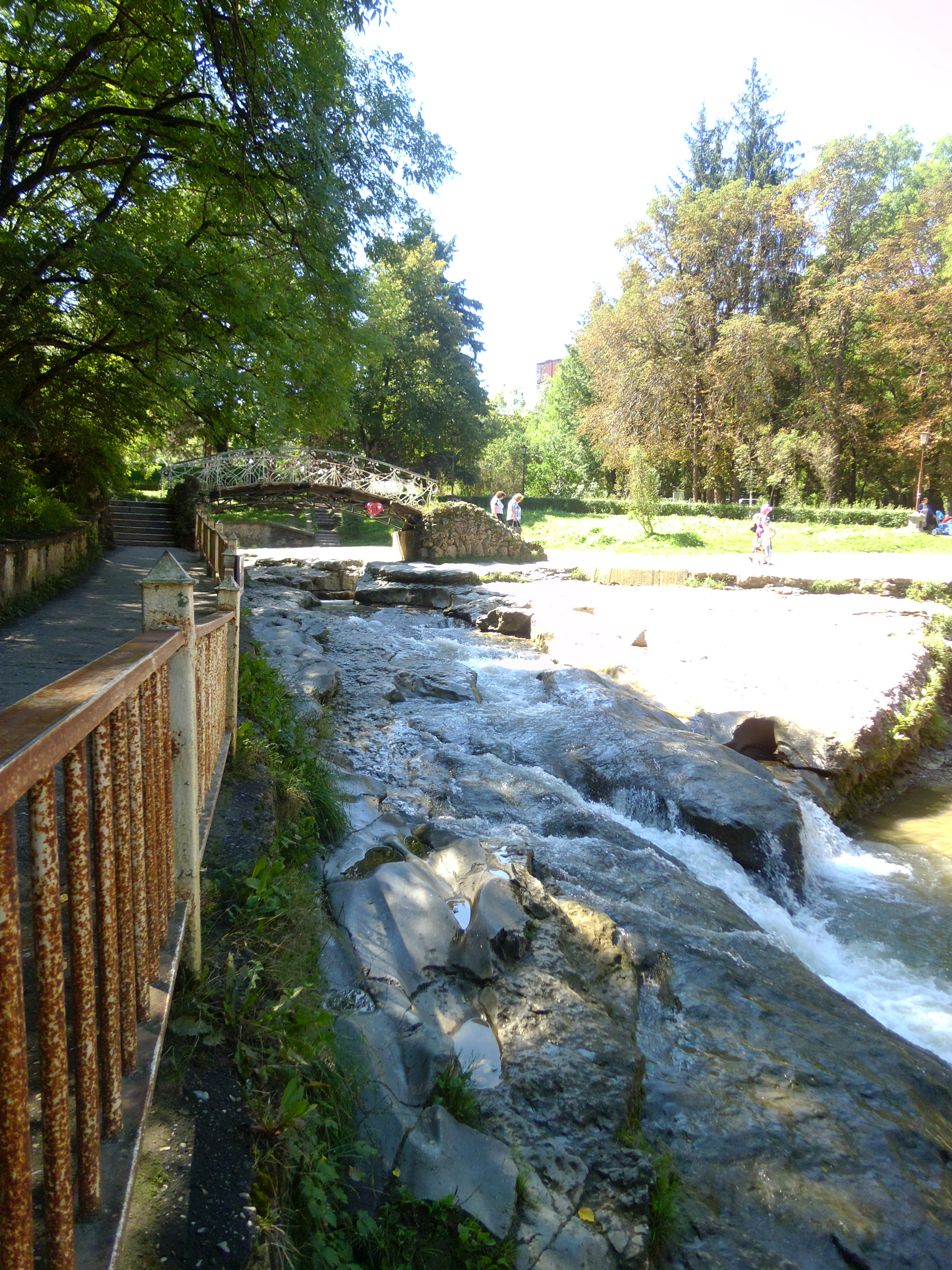 A stone arched bridge called Damsky Kapriz crossing a rocky, shallow river in a lush park setting.