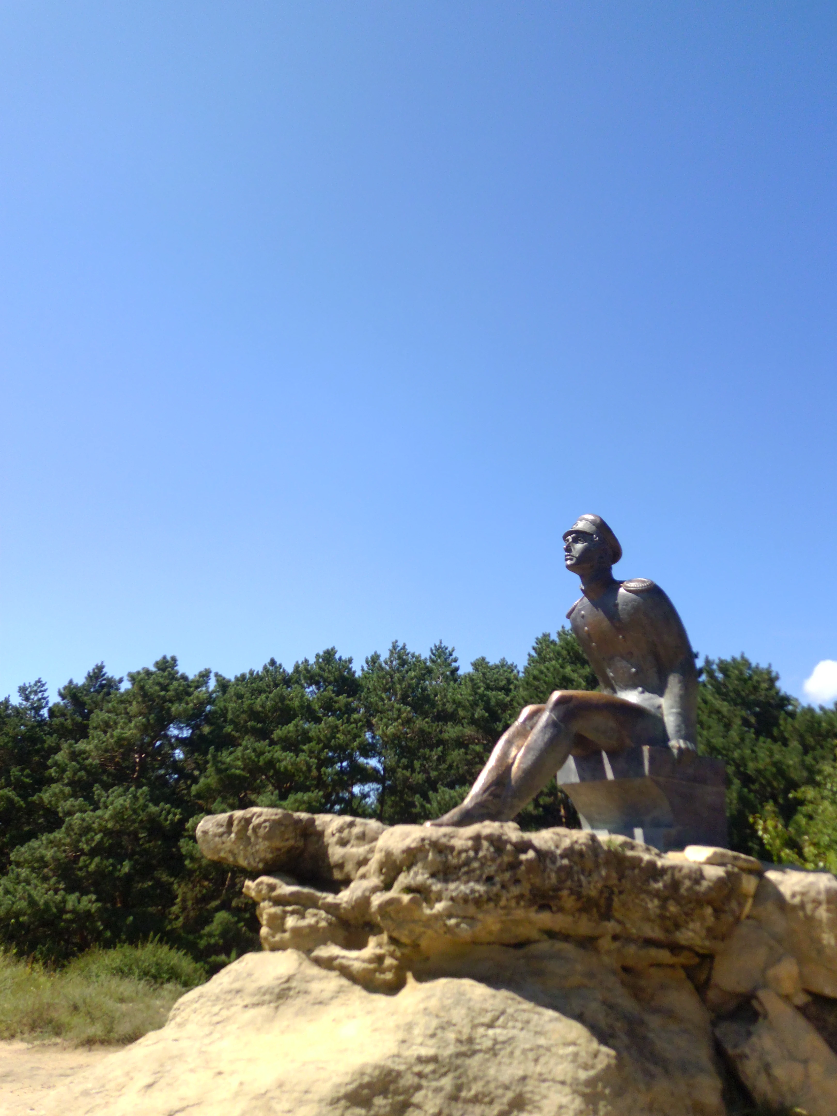 A bronze statue of a man in military uniform sitting on a rocky outcropping against a clear blue sky, with a forest in the background.