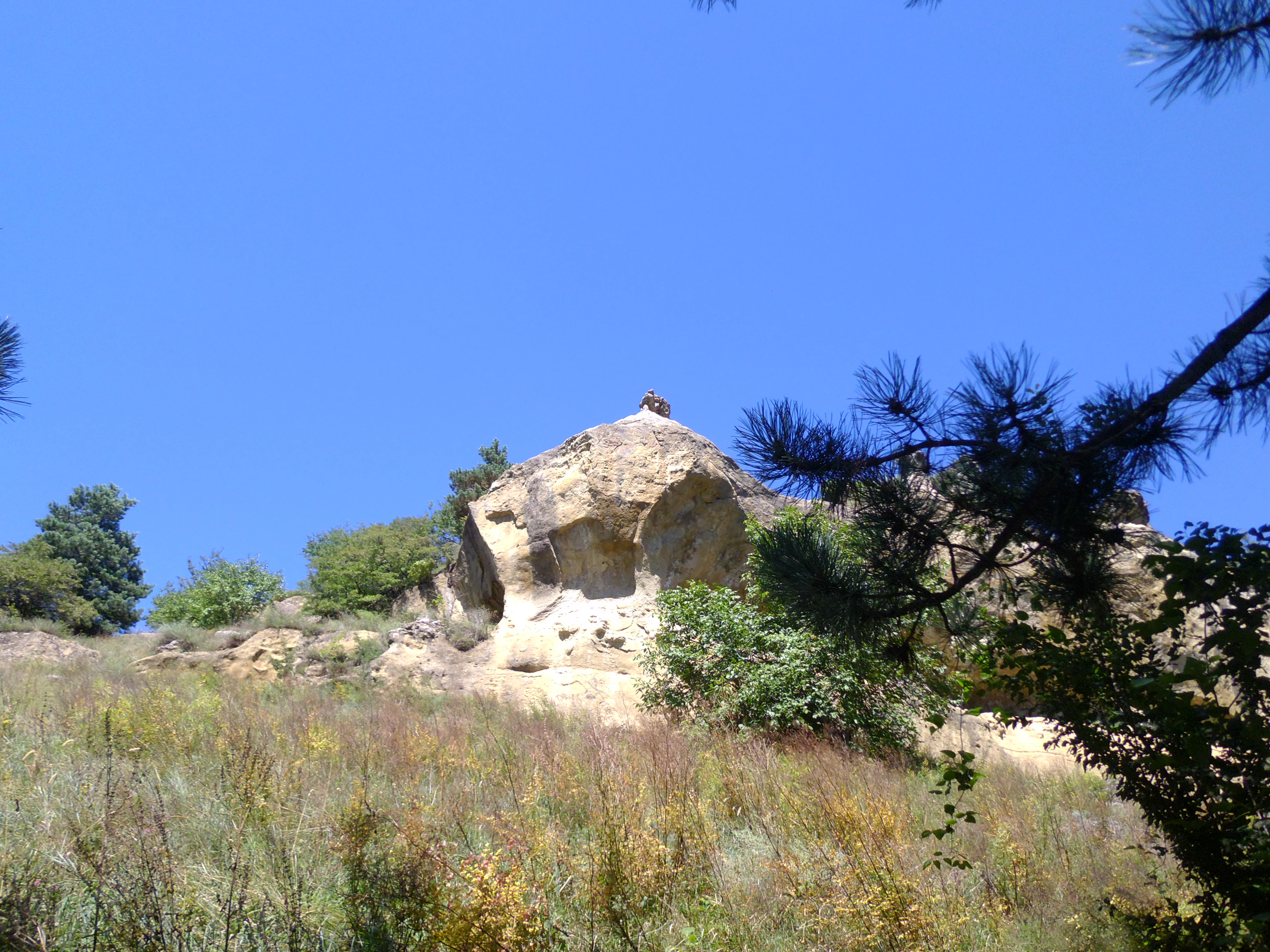 A low angle view of a light-colored, weathered sandstone rock formation rising above a grassy, vegetated slope against a bright blue sky.