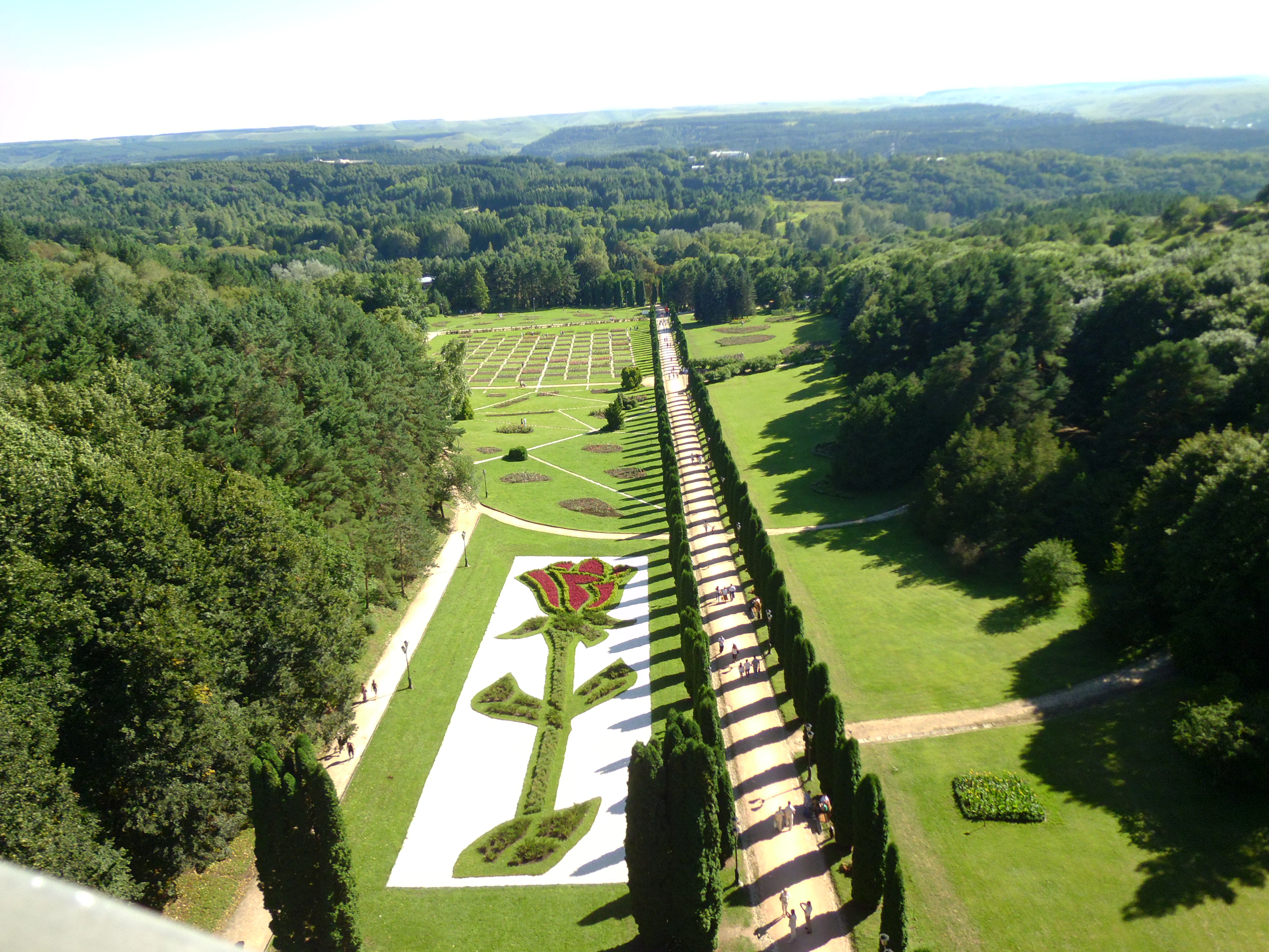 Aerial view showing a large garden with a rose-shaped floral design, surrounded by green fields, trees, and pathways with people walking
