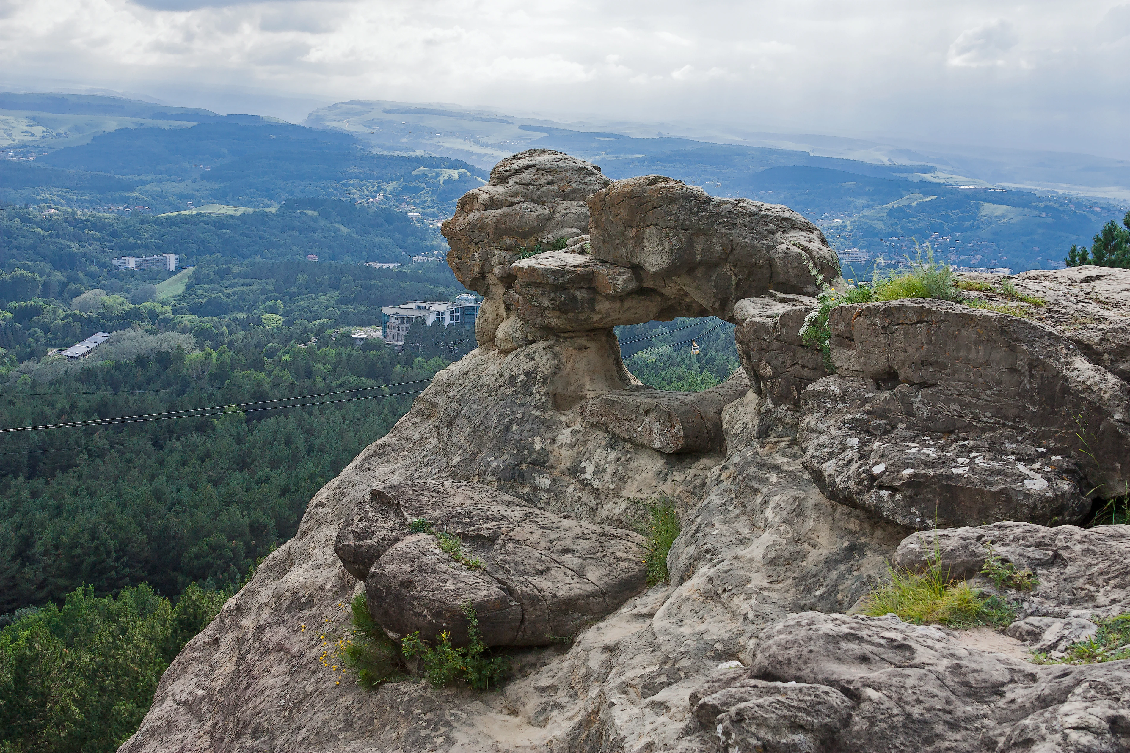 Rock arch formation on a hillside with forested valley and distant city under cloudy sky