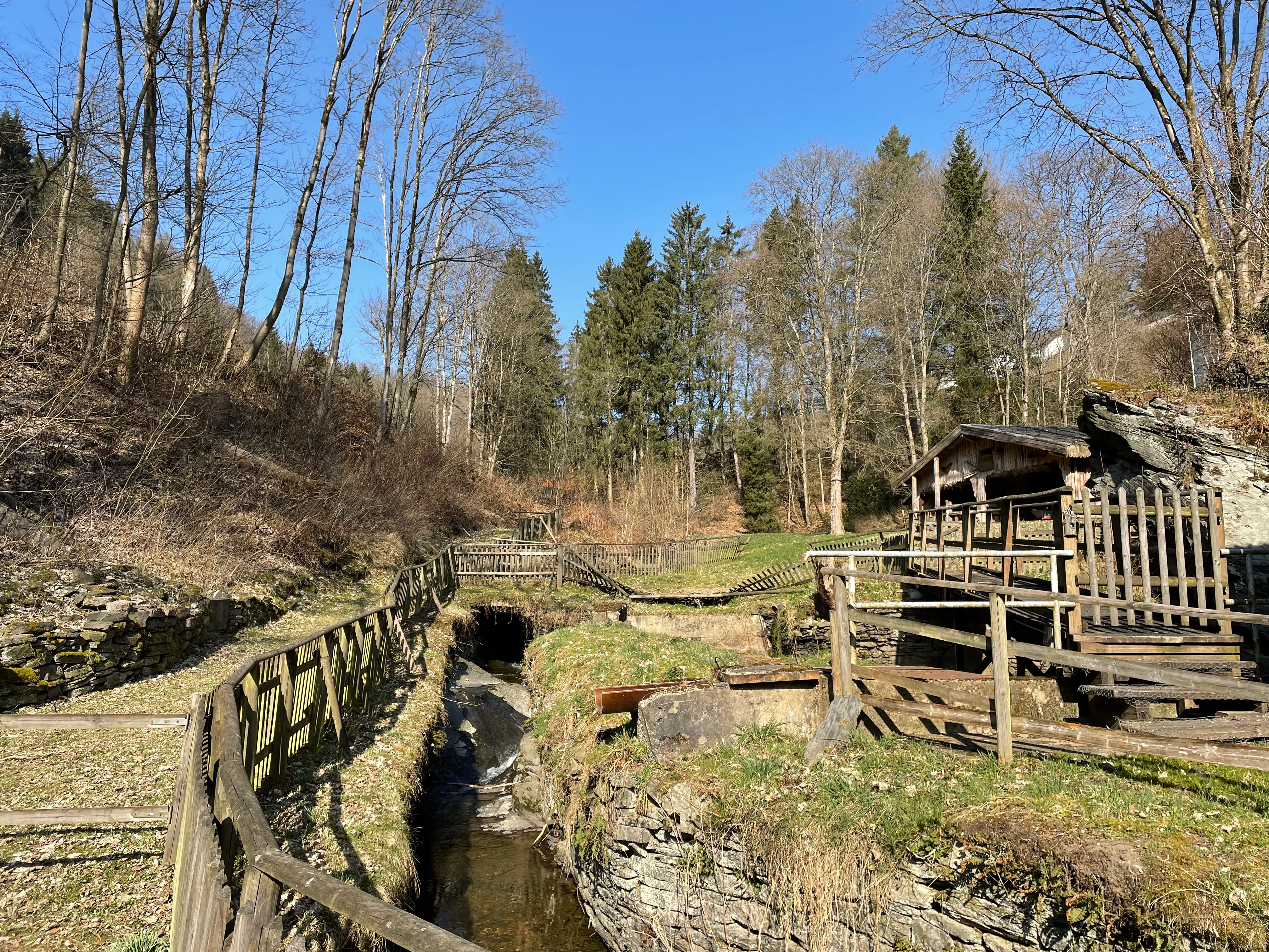 A clear, sunny day view of a small stream flowing through a valley, flanked by wooden fences and surrounded by bare deciduous trees and evergreen pines.