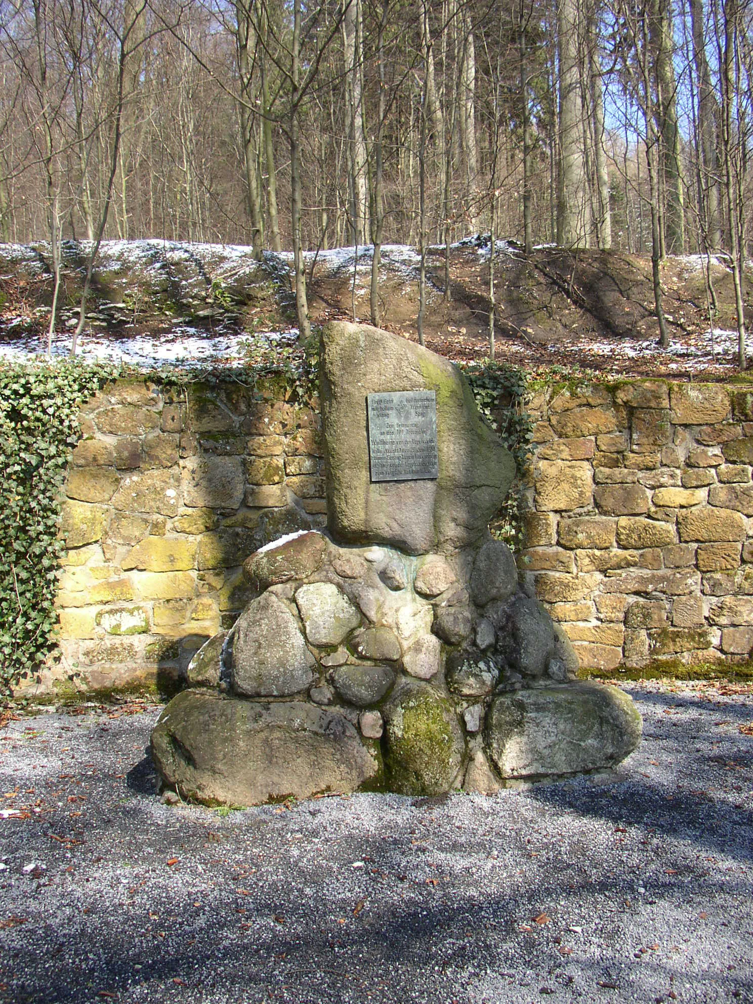 A stone monument built from stacked boulders with a metal plaque, situated in front of a stone retaining wall and a forest background with patches of snow.
