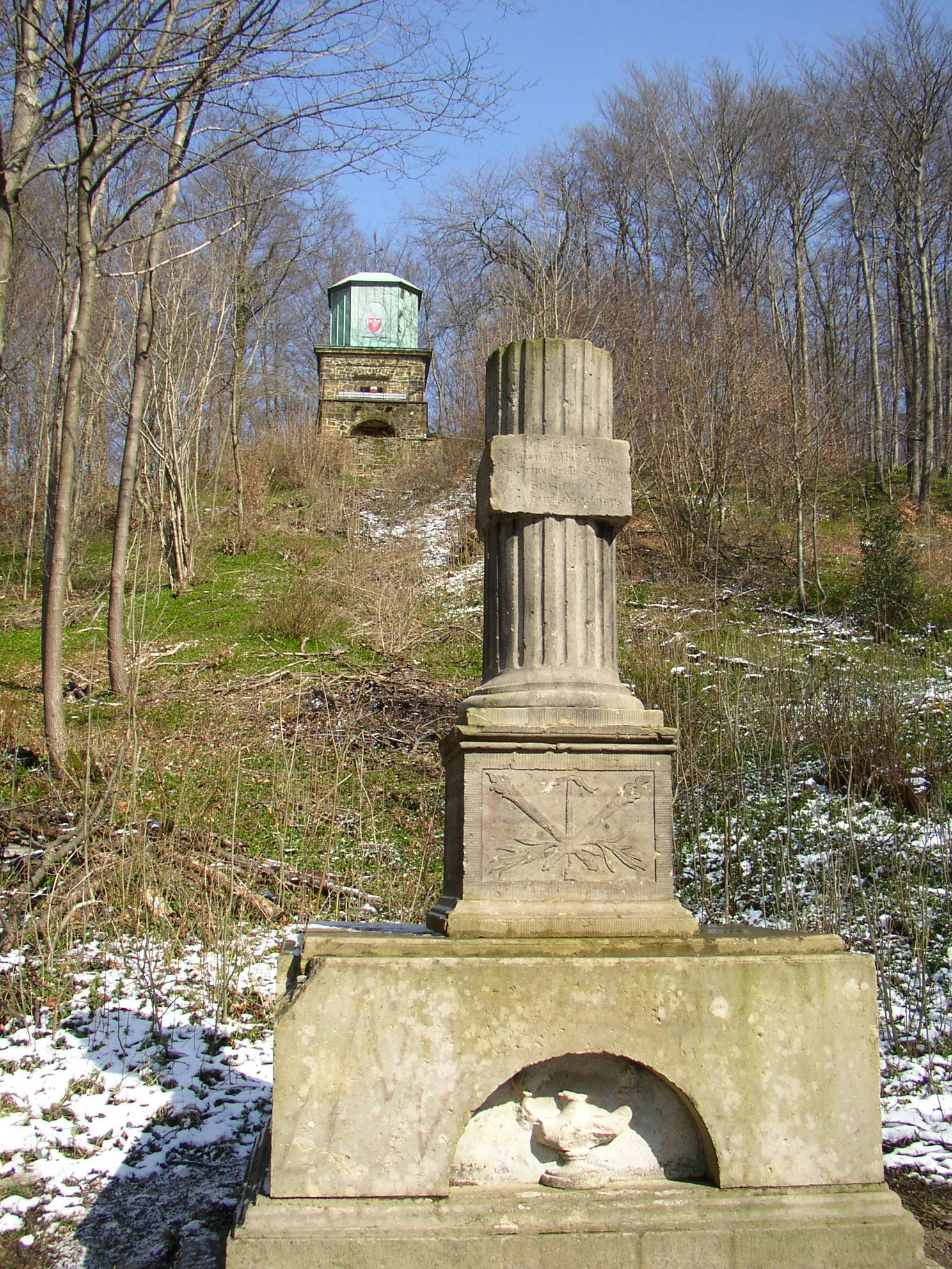 A stone monument with a fluted column stands in the foreground on a gentle, partially snow-covered slope, with a small stone tower, known as the Kaffeemühle, visible in the distance among bare trees.
