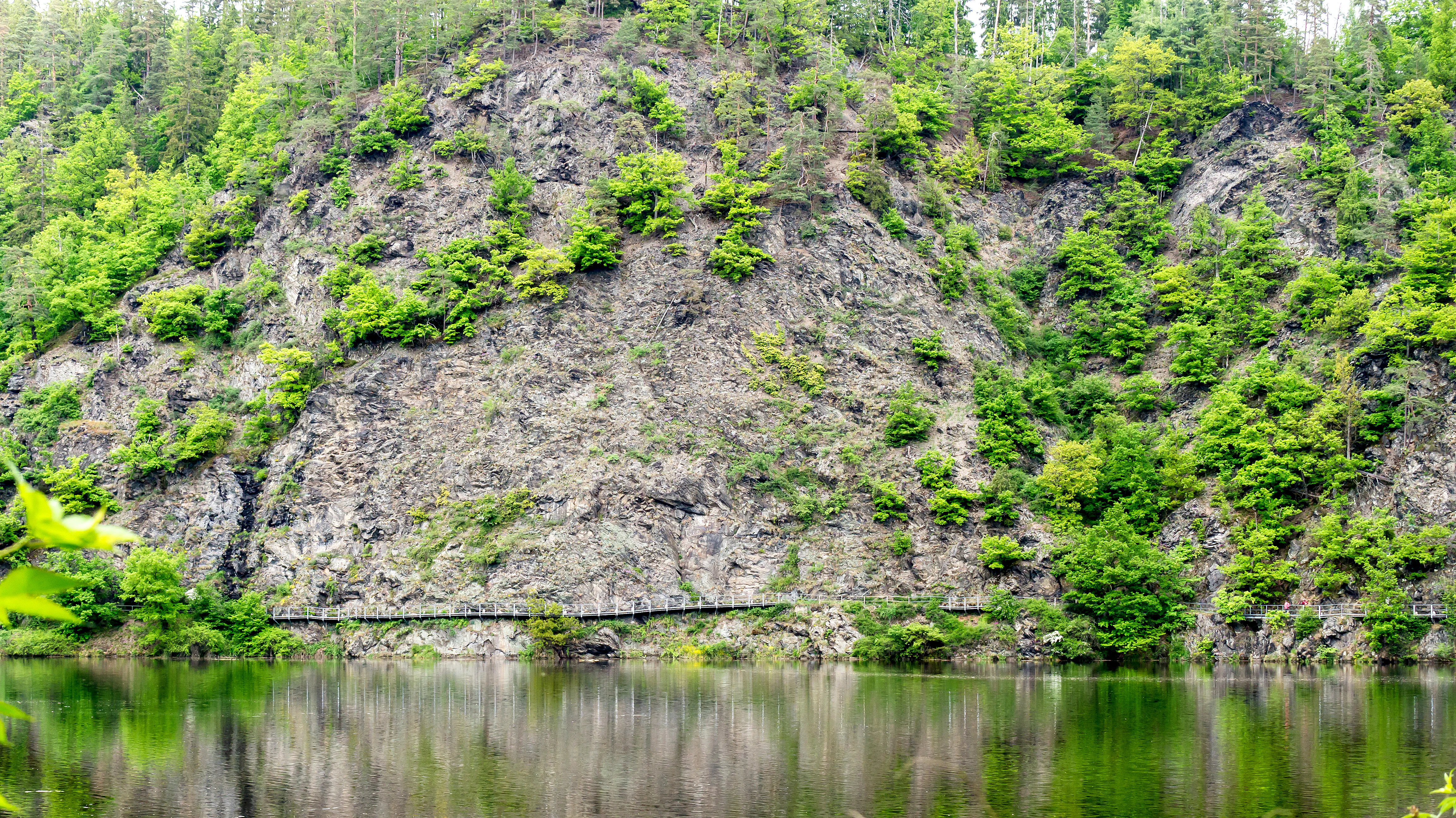A steep, rocky cliff face covered in sparse greenery, reflecting into the calm water of a reservoir at the base.