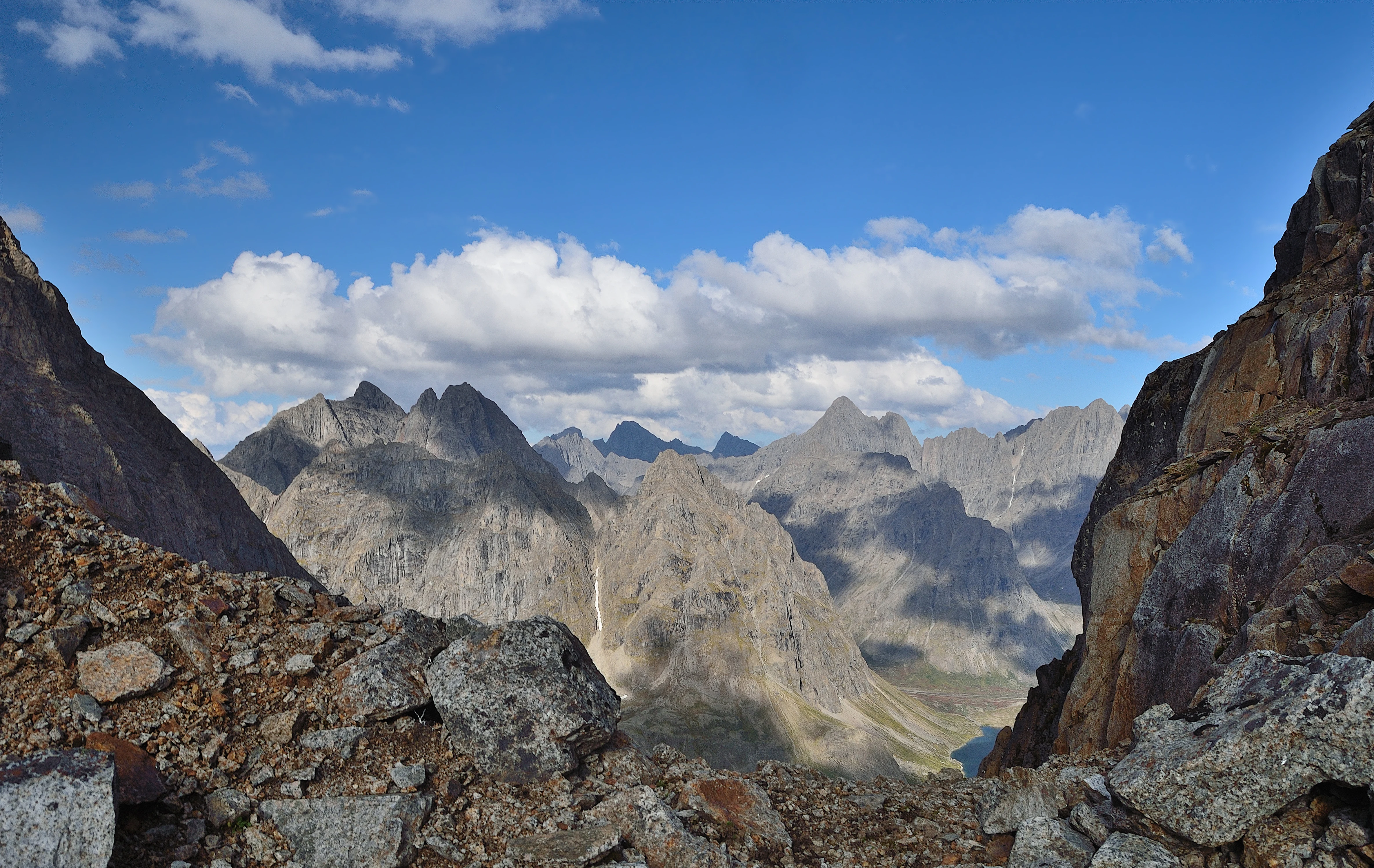 Rocky mountain pass with scattered stones in foreground, distant peaks under blue sky with clouds, and Uglovoe Lake visible in valley