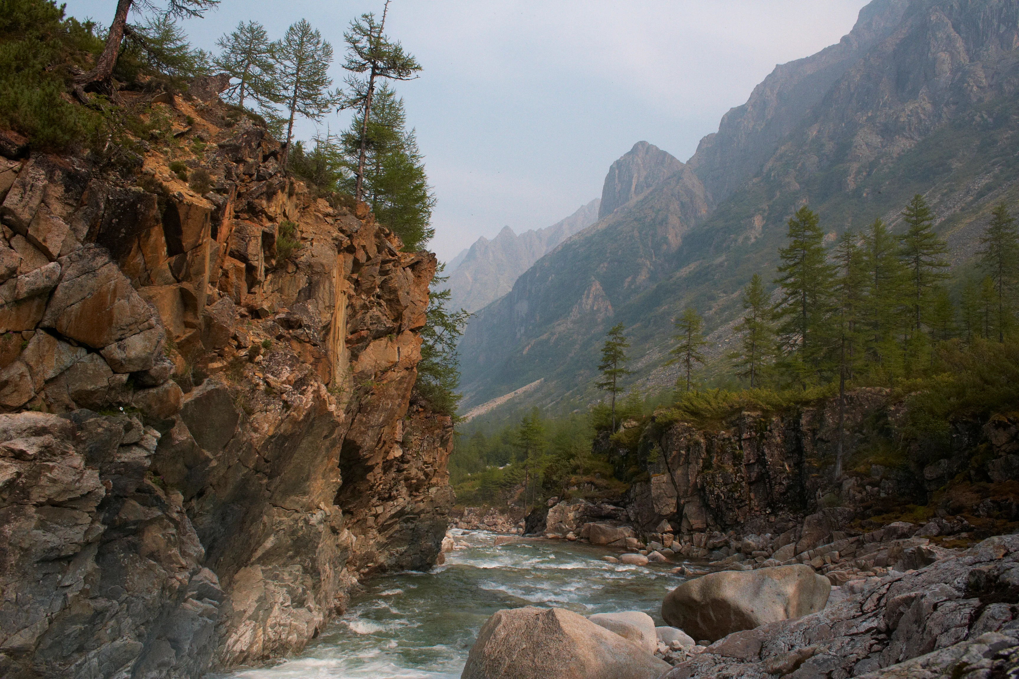 A river flowing through a rocky valley with steep cliffs and coniferous trees, surrounded by mountain peaks under a partly cloudy sky
