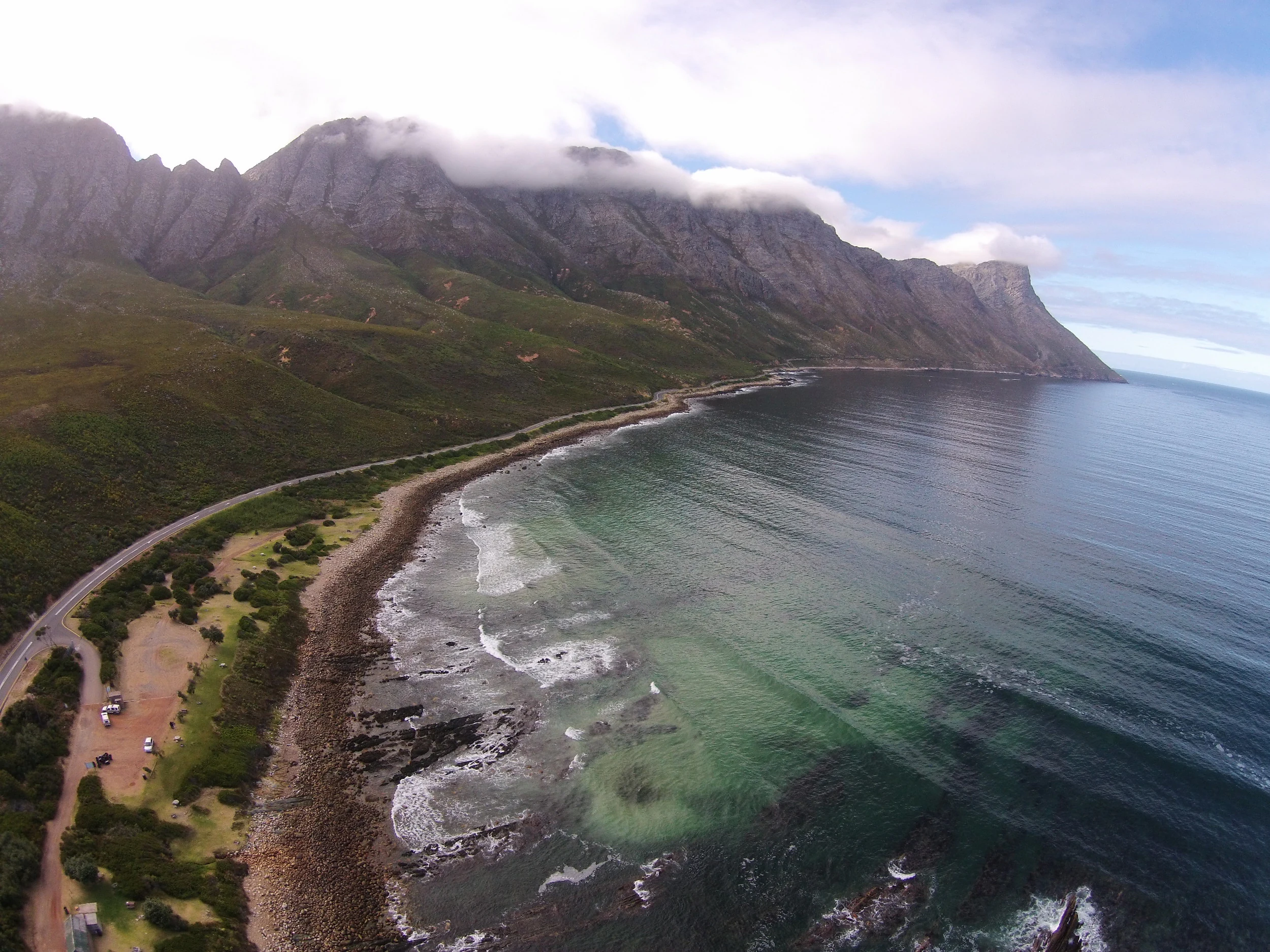 An aerial view of a coastline with a long beach and rocky shore, a winding coastal road, and high mountain ridges partially obscured by clouds.