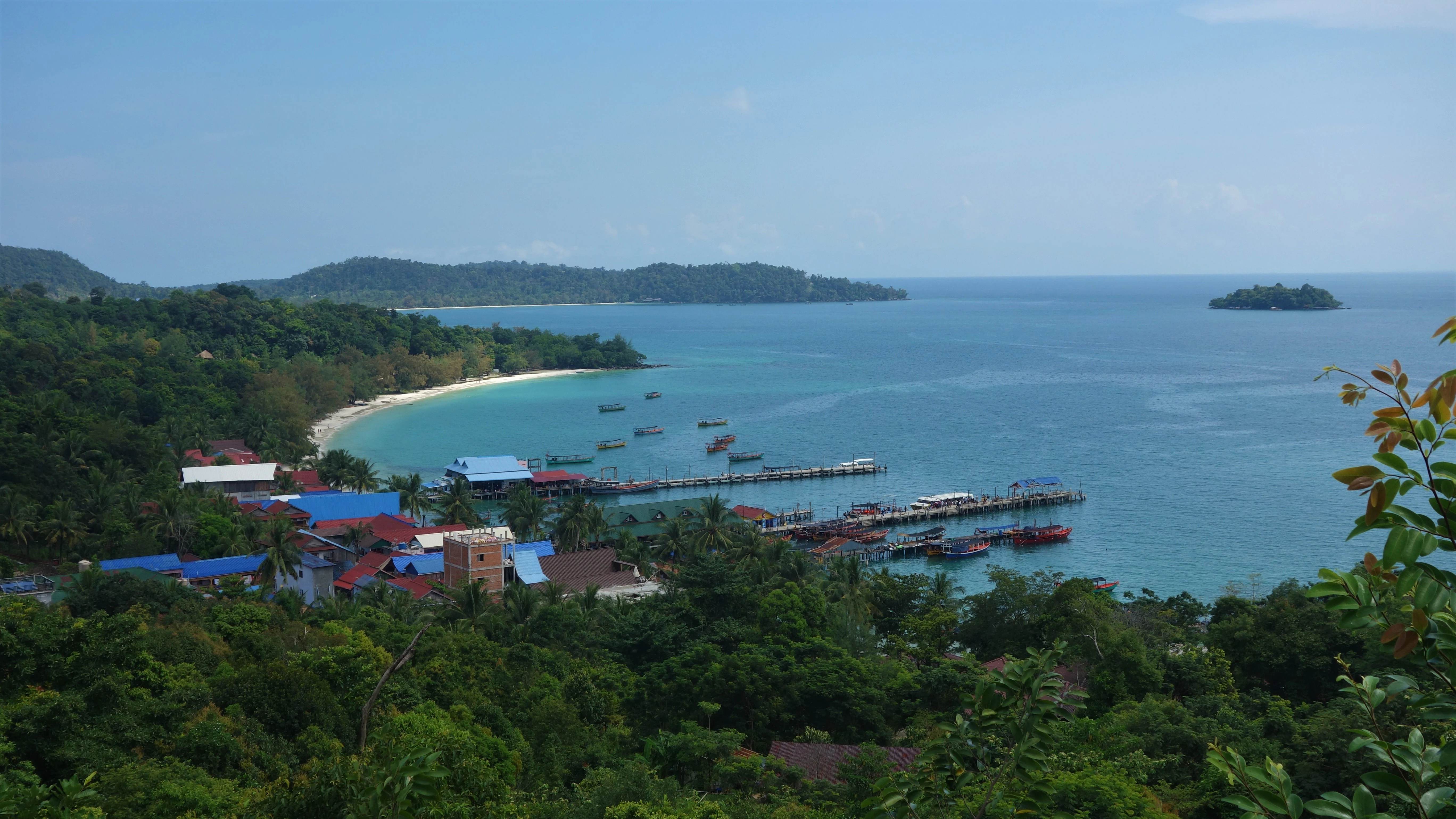 Panoramic coastal landscape showing sandy beach, turquoise ocean, docked boats, and dense green forest with buildings along the shoreline