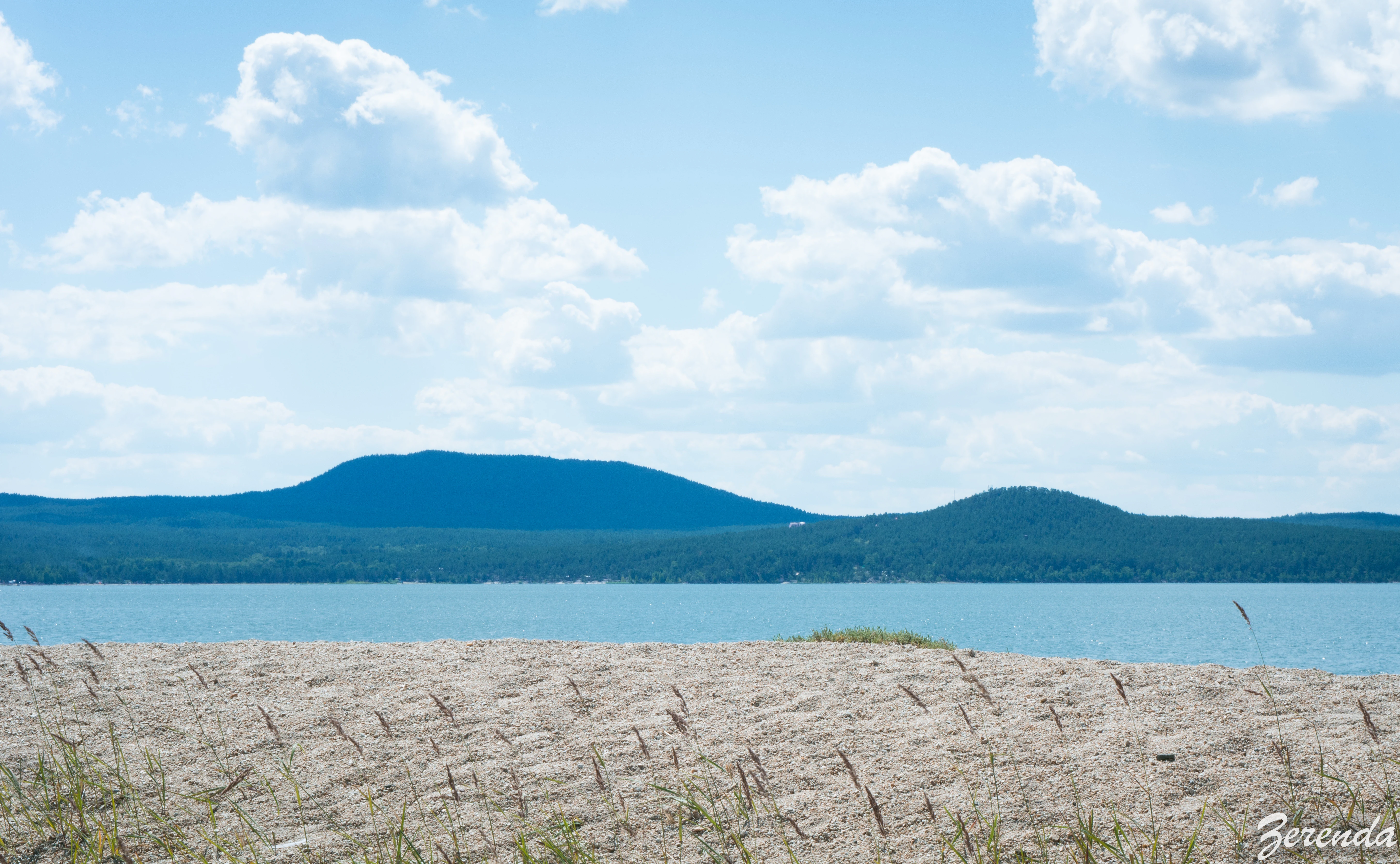 Sandy beach with sparse grass, a body of water, and distant blue mountains under a partly cloudy sky