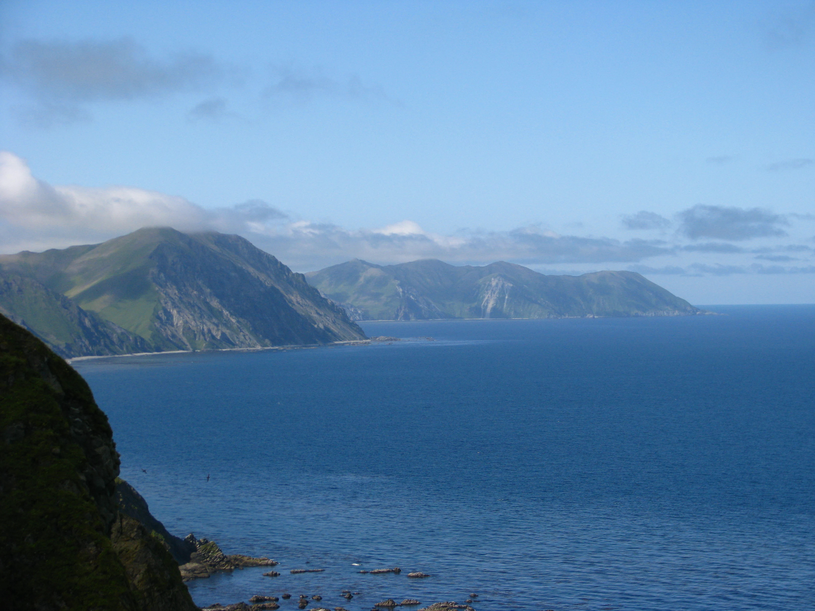 A wide angle view of a mountainous green island coastline meeting the blue ocean under a partly cloudy sky.