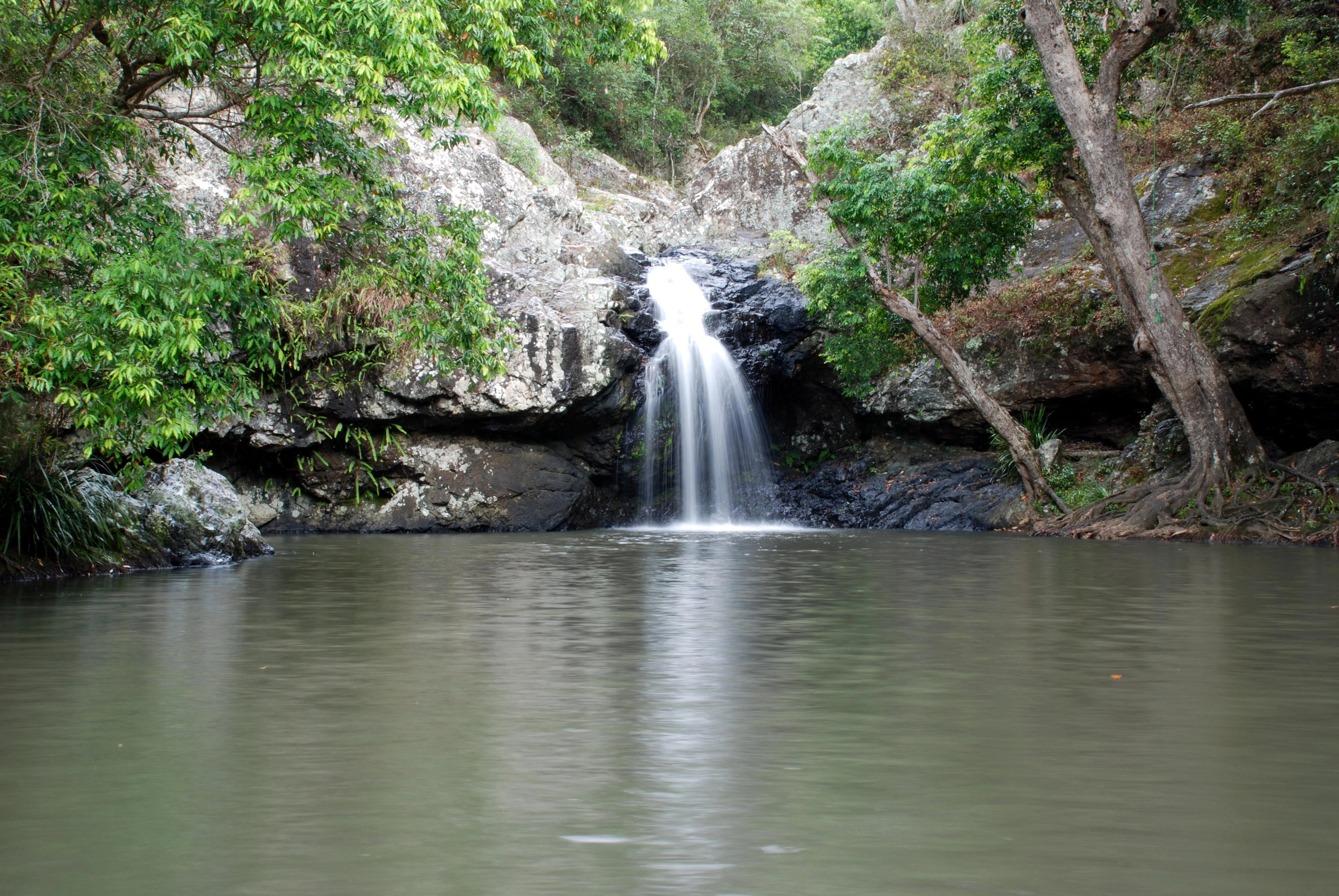Waterfall flowing into a calm pool surrounded by lush green trees and rocky cliffs.