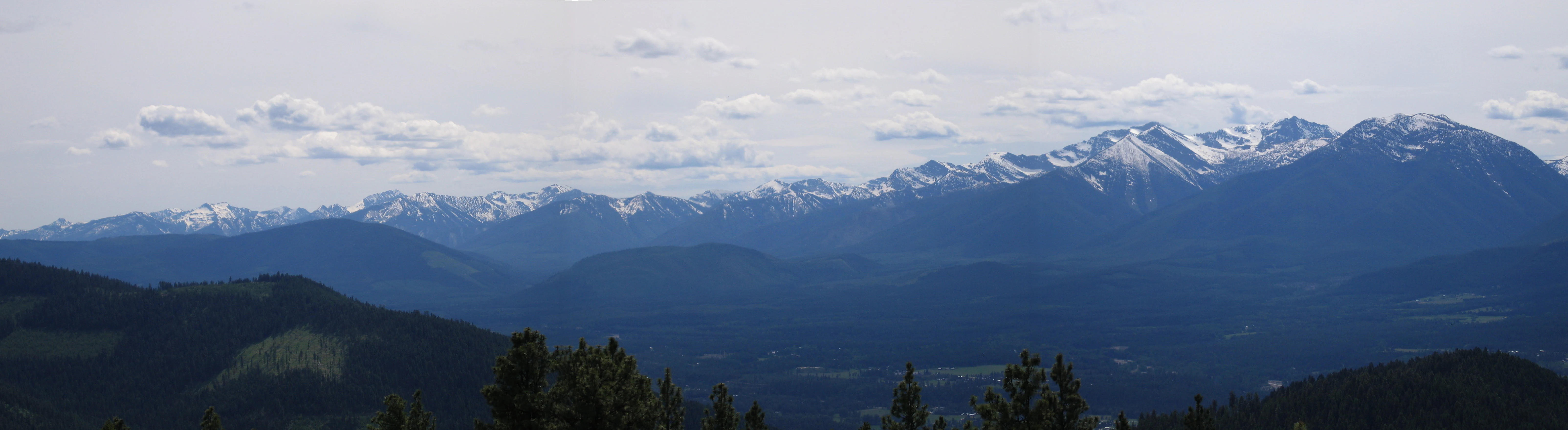 A wide panoramic photograph showing a rugged mountain range with patches of snow on the peaks, receding into layers of forested ridges under a cloudy sky.