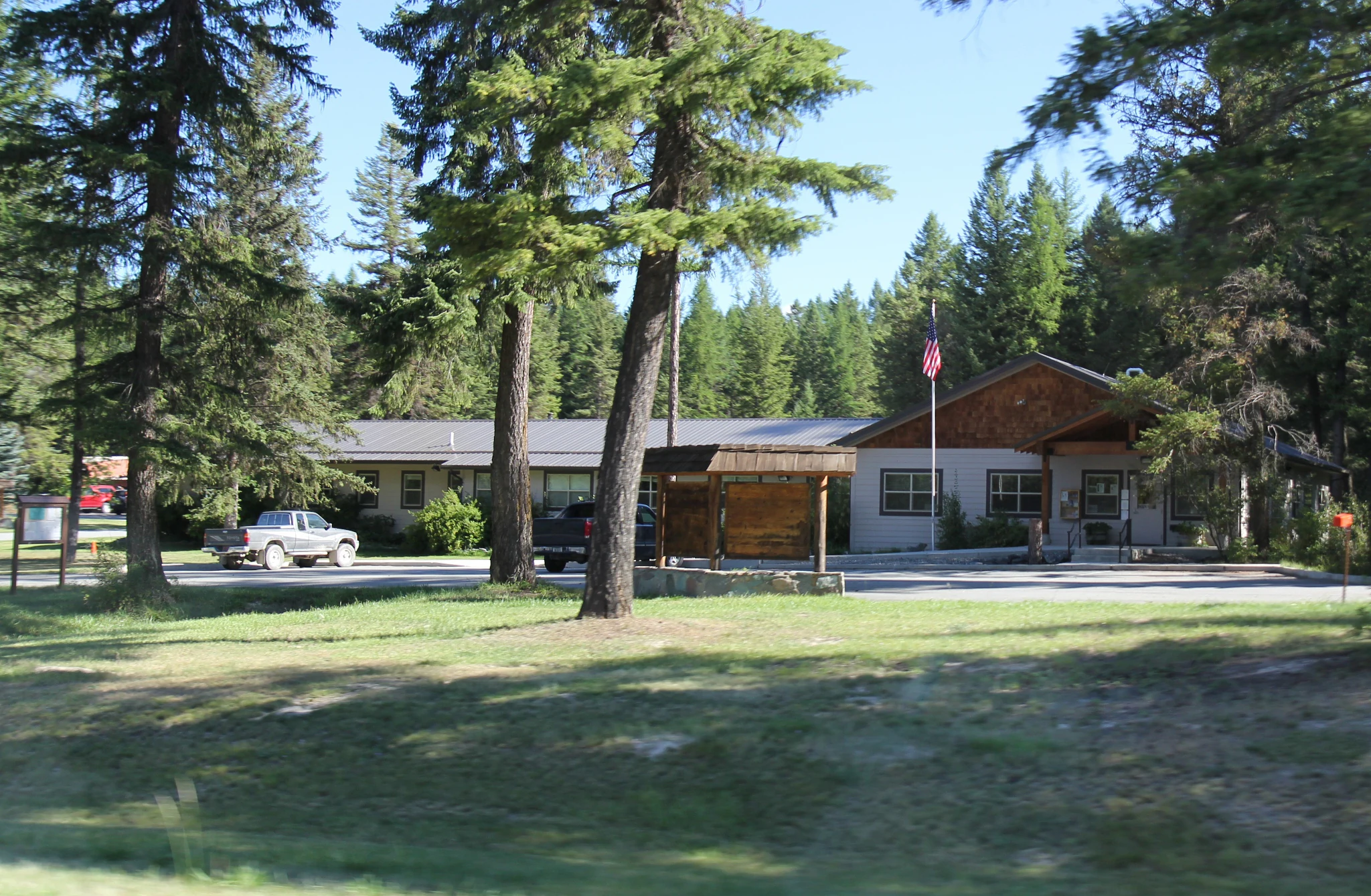 A one-story building serving as a ranger station, surrounded by tall pine trees with a parking area in front containing a pickup truck.