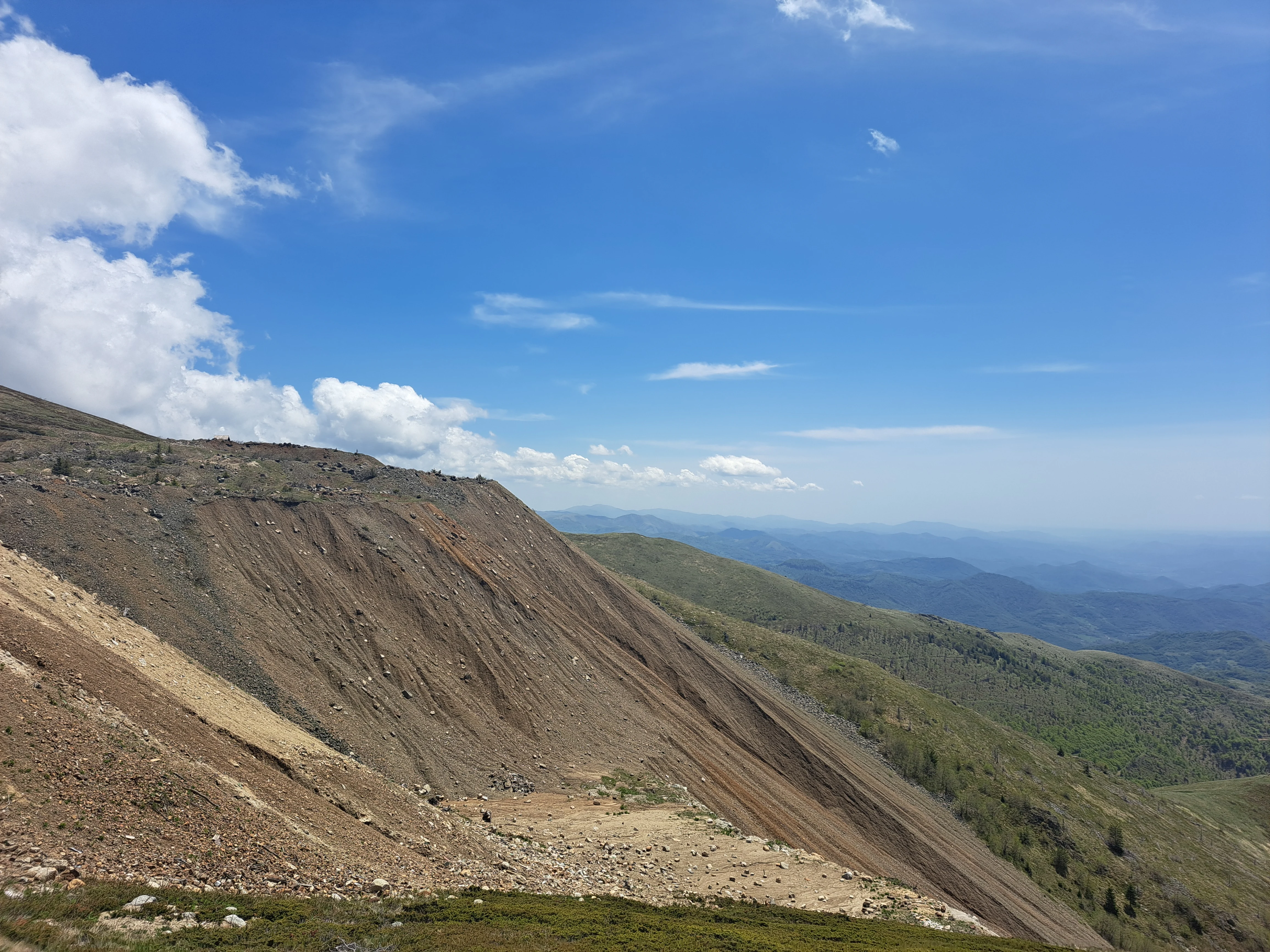 Rocky mountain slope with patches of green vegetation, distant hills, and a blue sky with scattered clouds