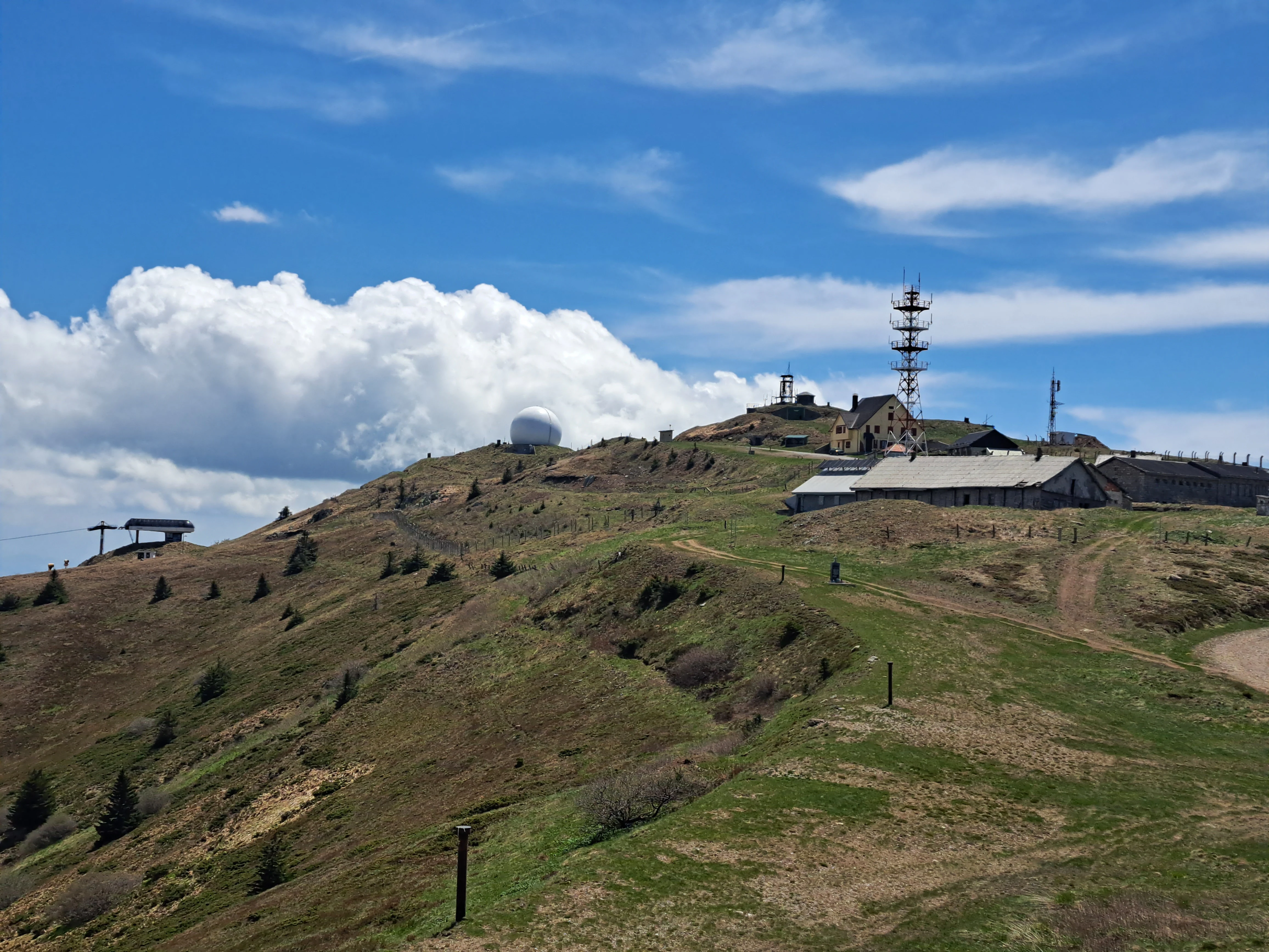 Green and brown mountain slopes with a building, communication tower, and clear blue sky with clouds