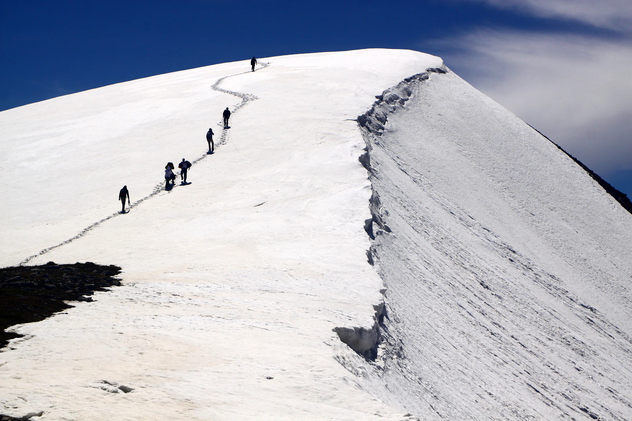 A group of hikers walking single file up a steep, snow covered mountain ridge under a clear blue sky.