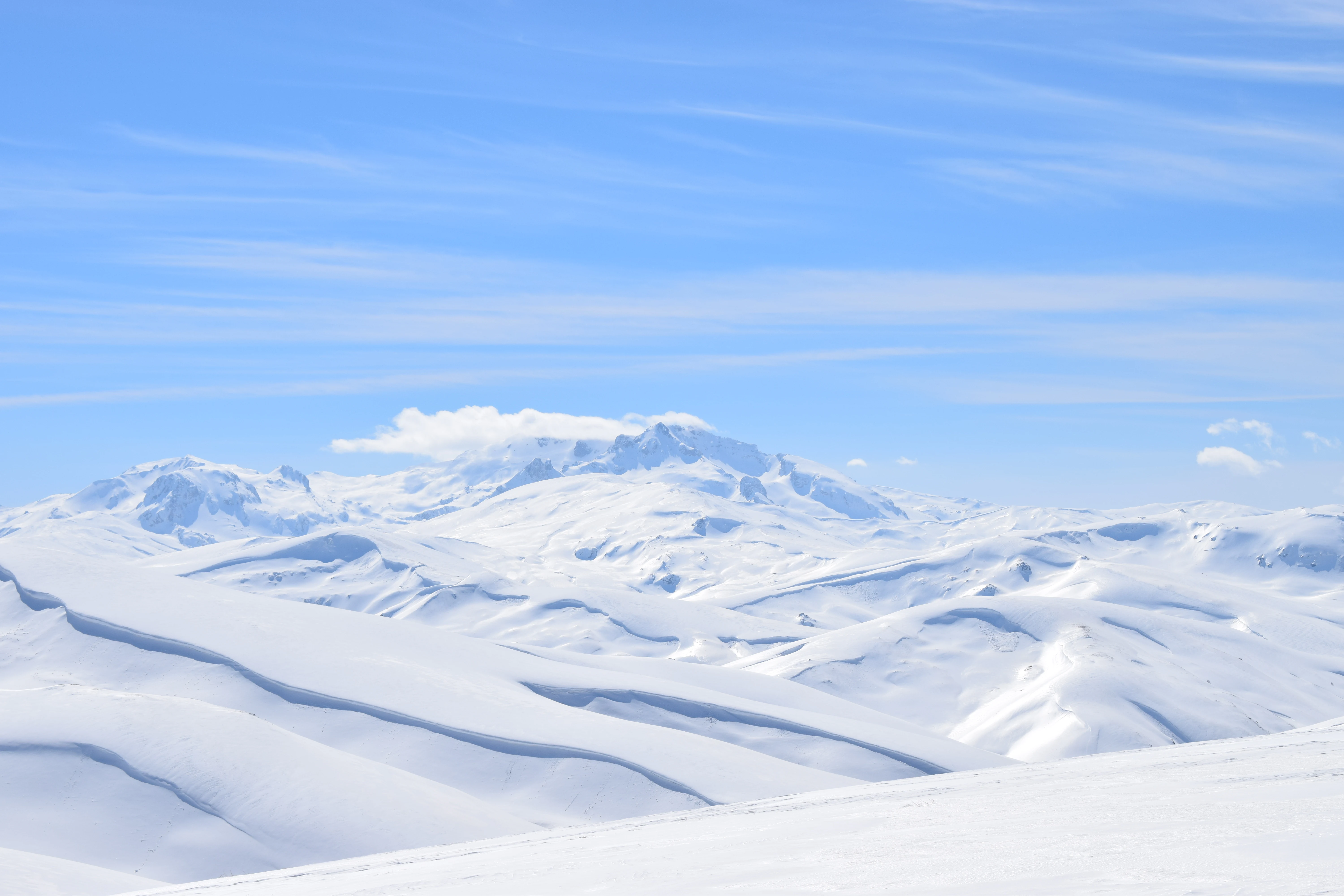 Wide shot of a vast, high altitude mountain range completely covered in deep snow under a bright blue sky.