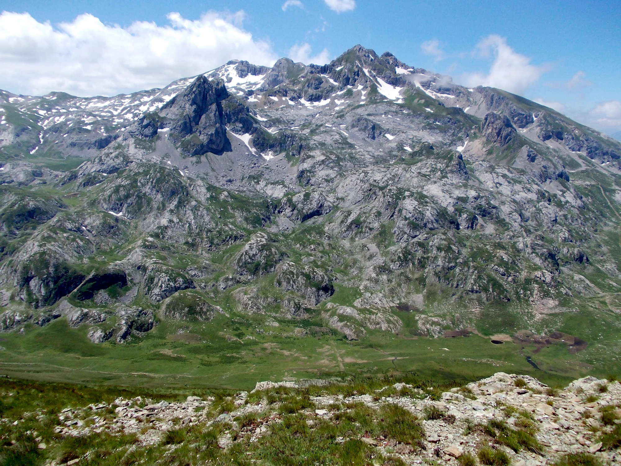 A wide view of a large, rocky mountain massif with patches of snow near the summit, rising above a green alpine valley.