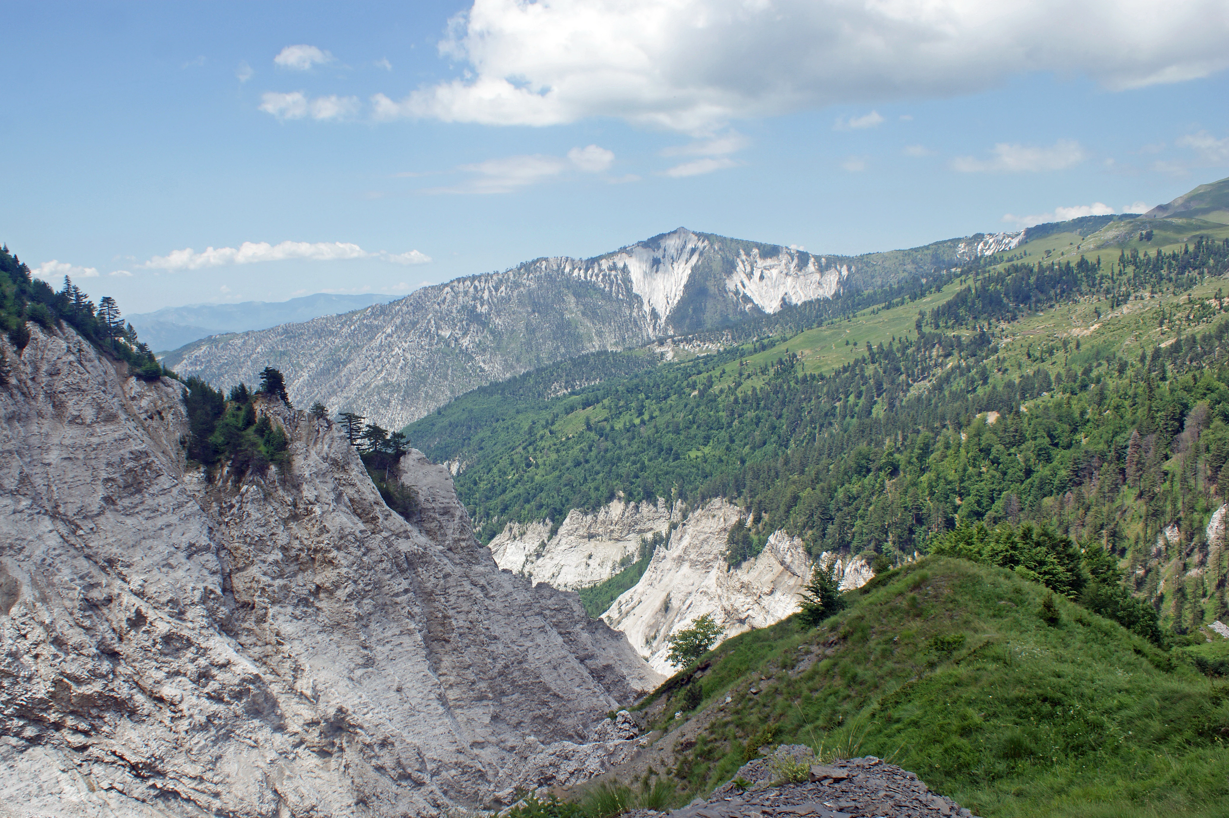 A mountainous landscape showing rocky slopes, forested hillsides, and a prominent peak under a partly cloudy sky.