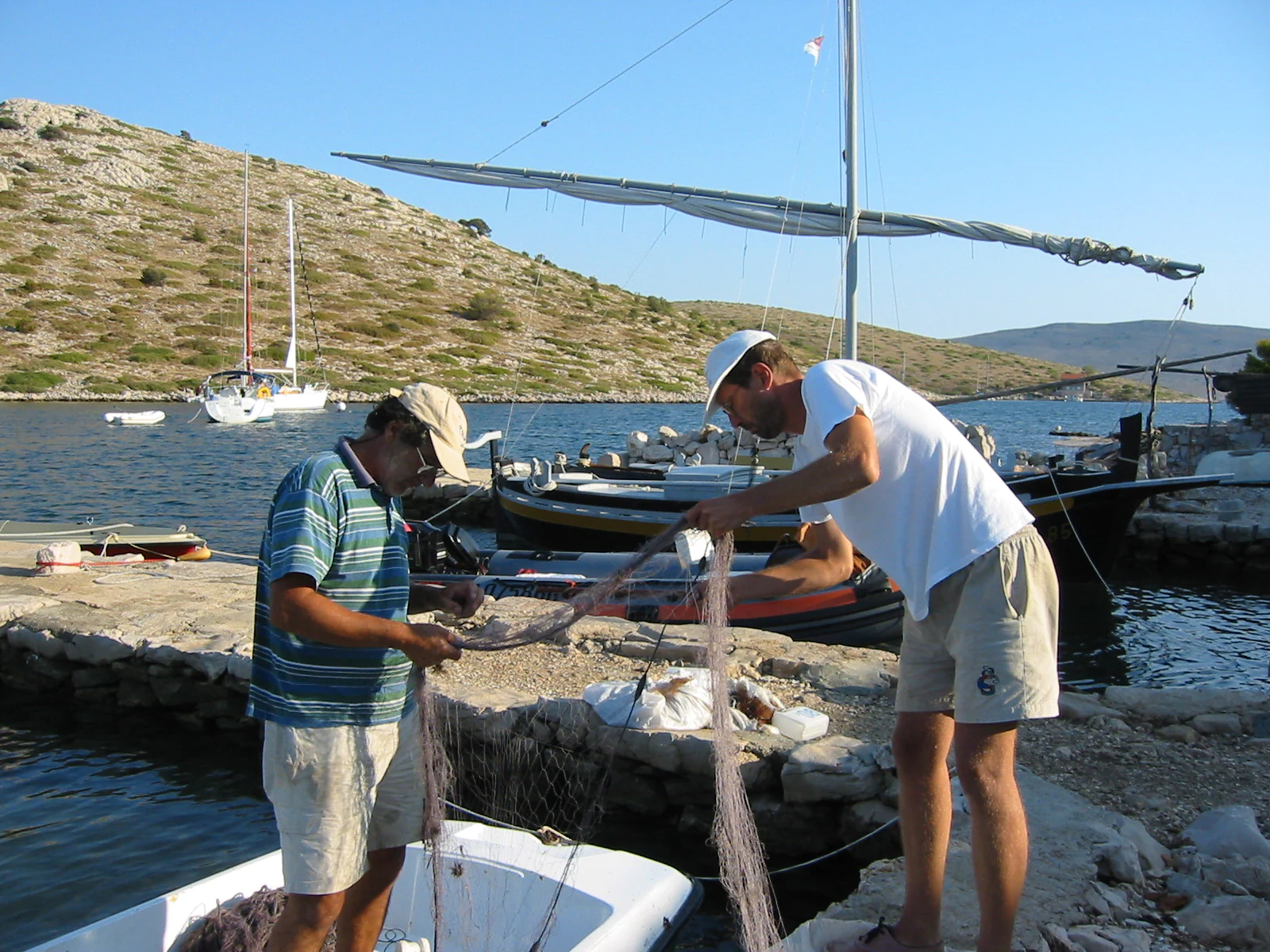 Two men handling fishing nets on a dock near boats and rocky coastline