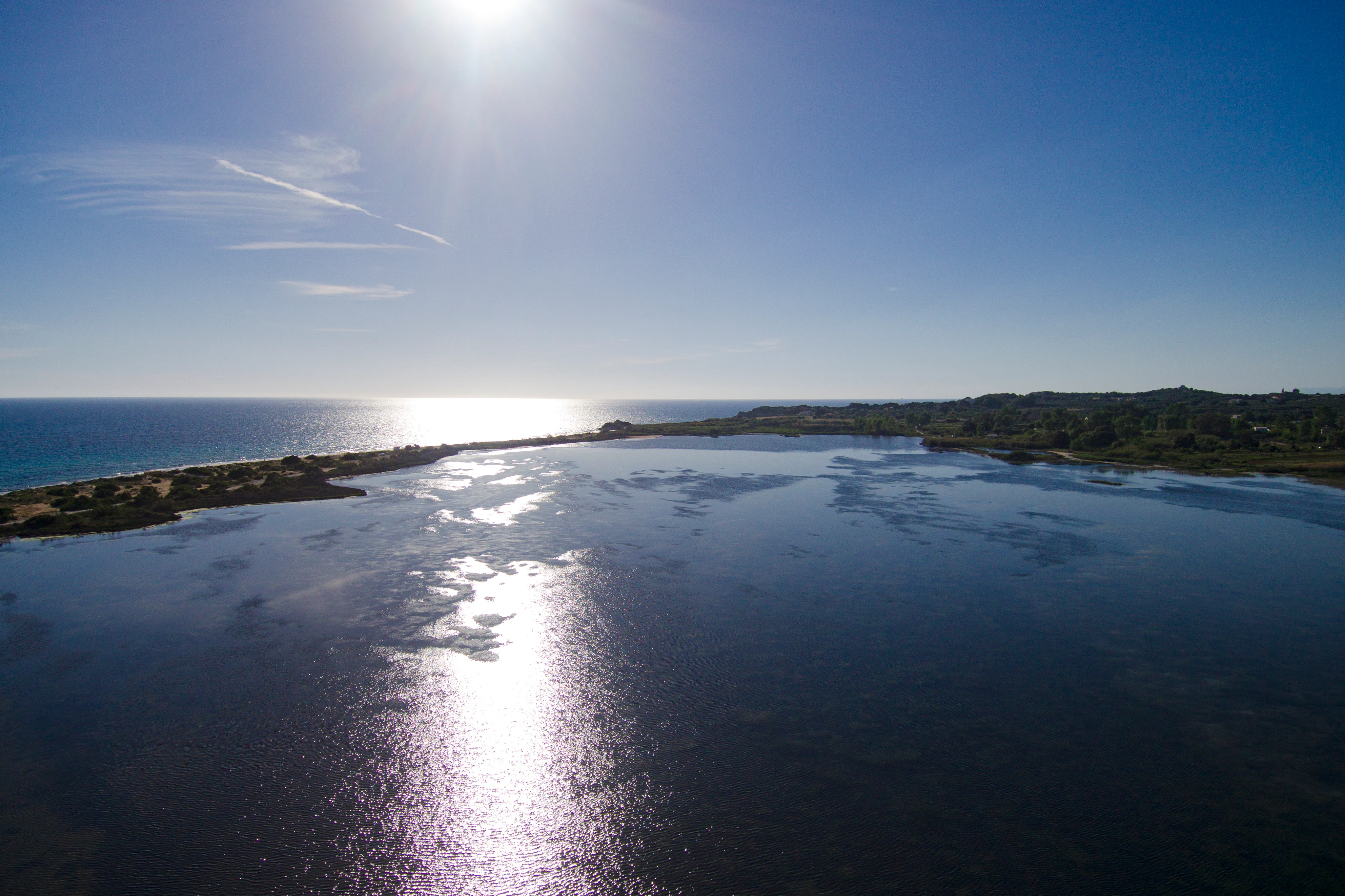 Aerial view of a coastal lagoon separated from the Ionian Sea by a narrow strip of land and sand dunes under a clear blue sky.