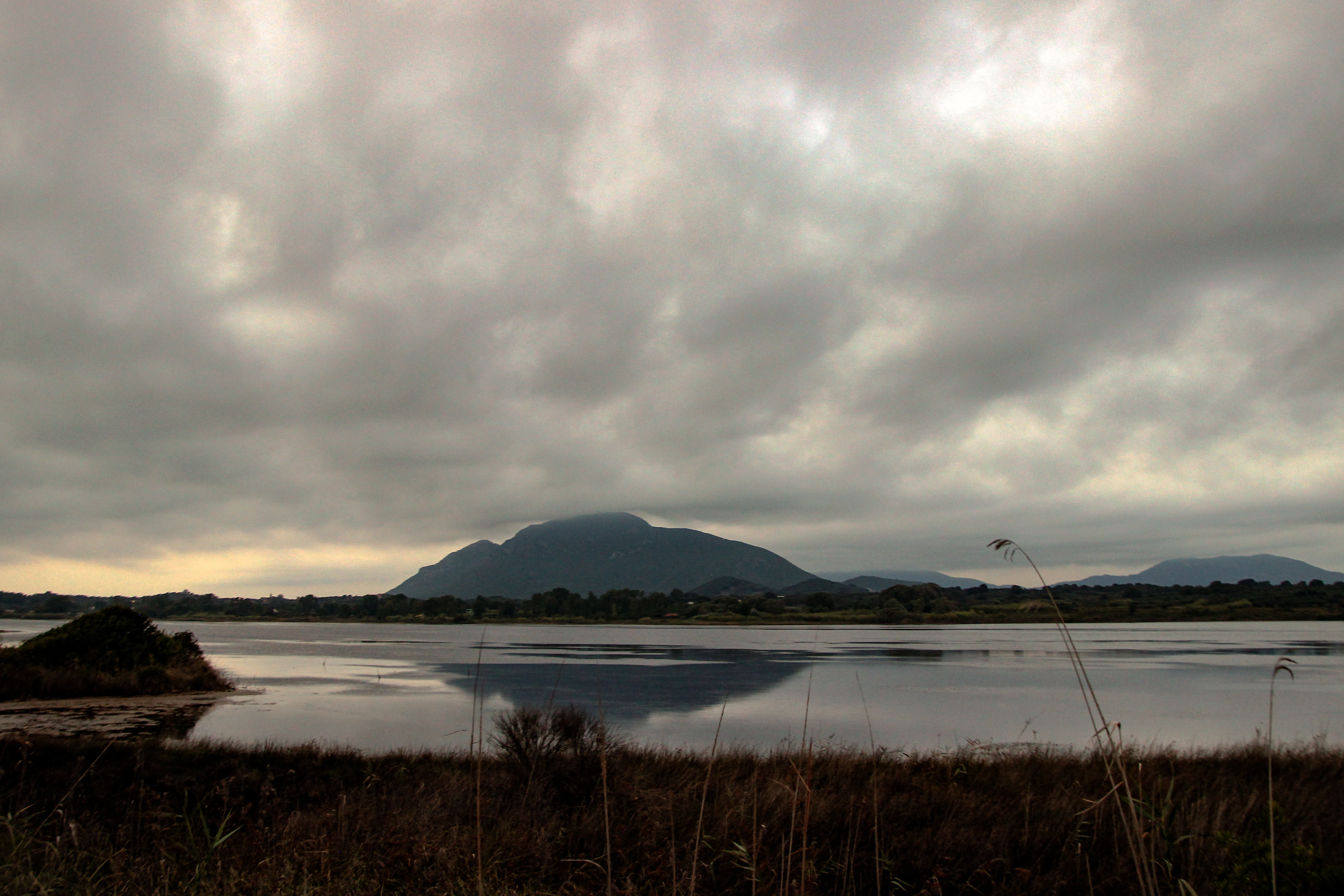 A wide landscape view of Korission Lagoon with a dark, prominent hill in the distance, mirrored in the calm water, under a heavy, overcast sky.