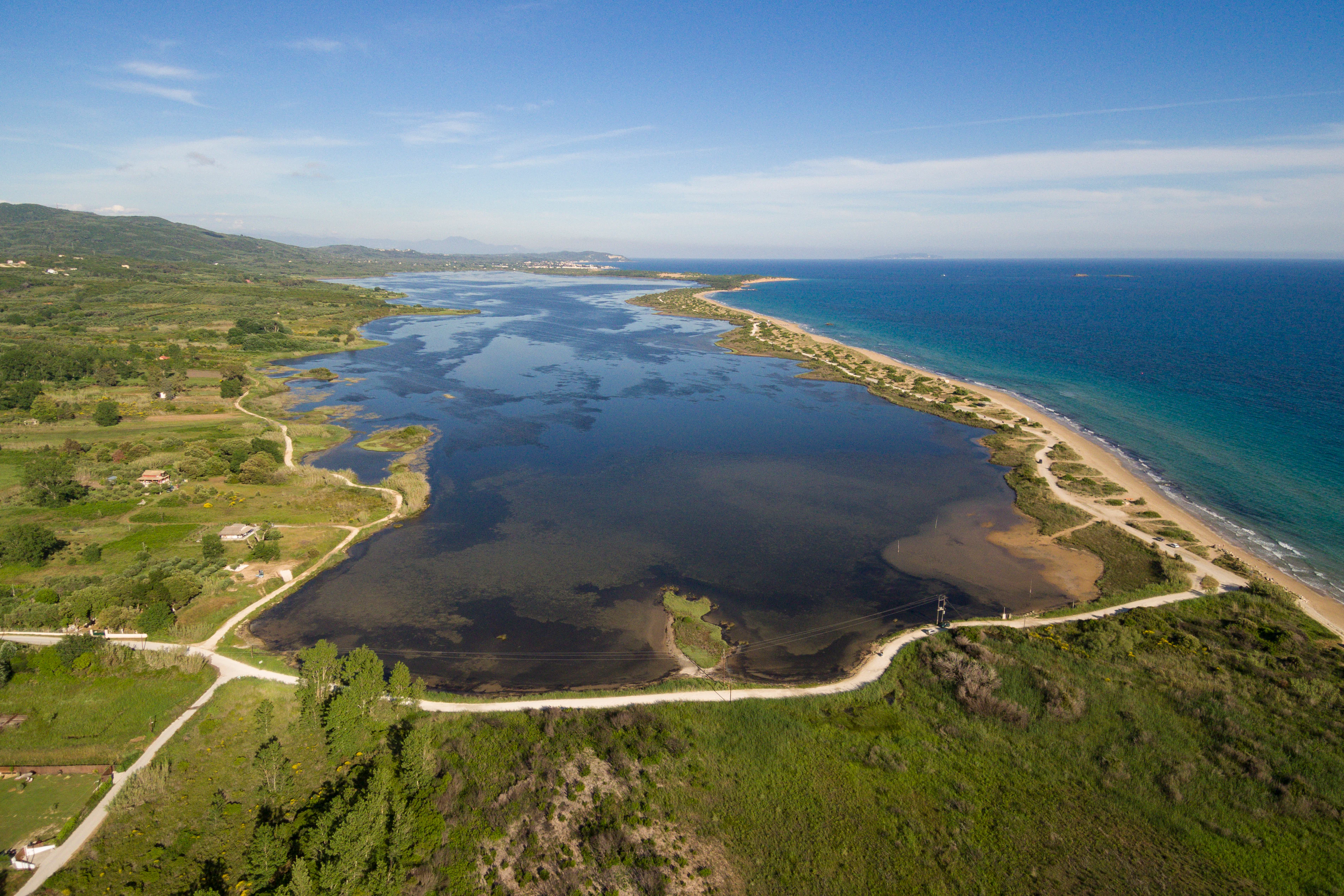 High-angle aerial photograph of Korission Lagoon in Corfu, Greece, with a large, dark-watered lagoon separated from the blue Ionian Sea by a thin, sandy coastal barrier and dunes.