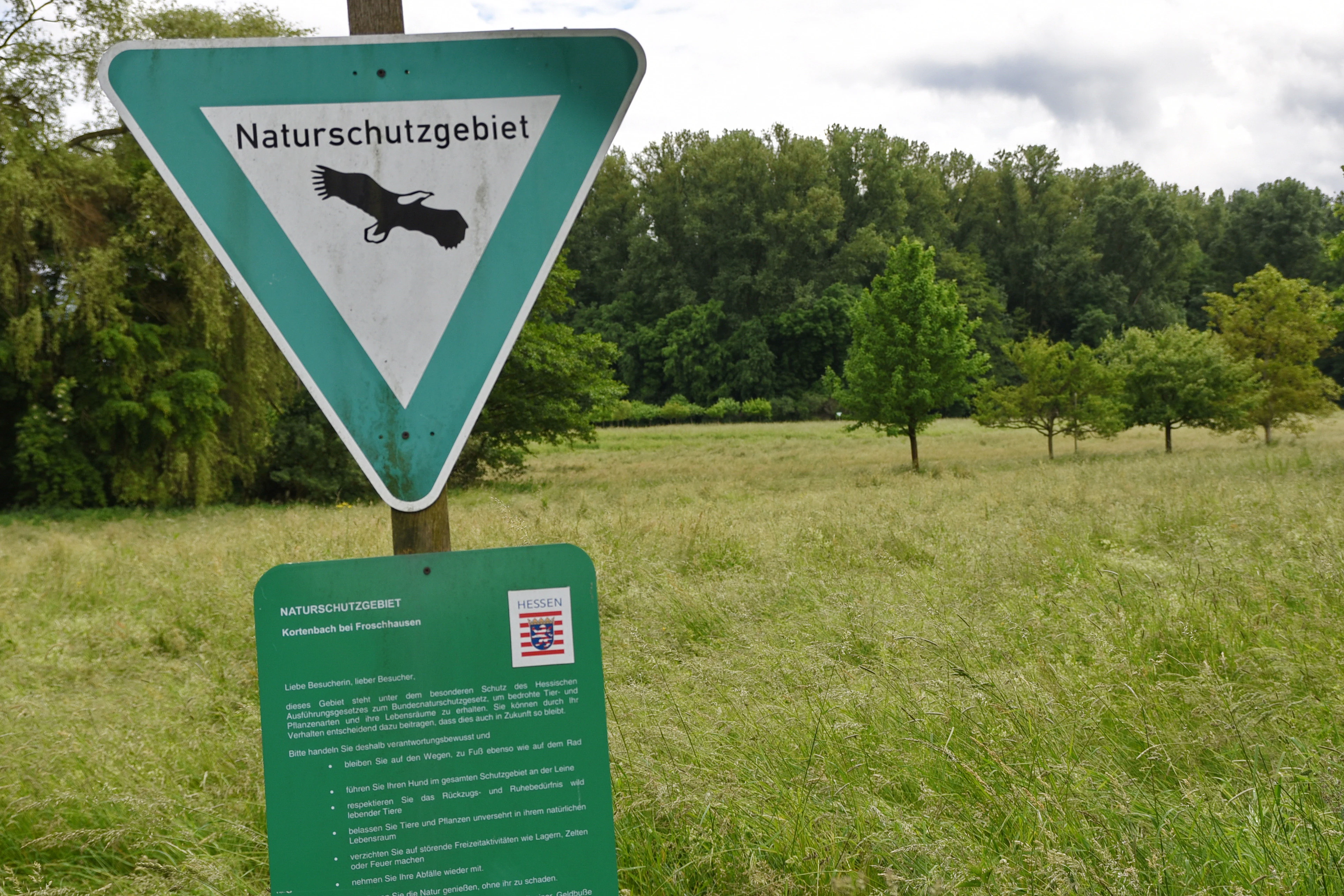 A triangular nature reserve sign with a bird silhouette and a green informational board below it, set against a grassy field and trees.