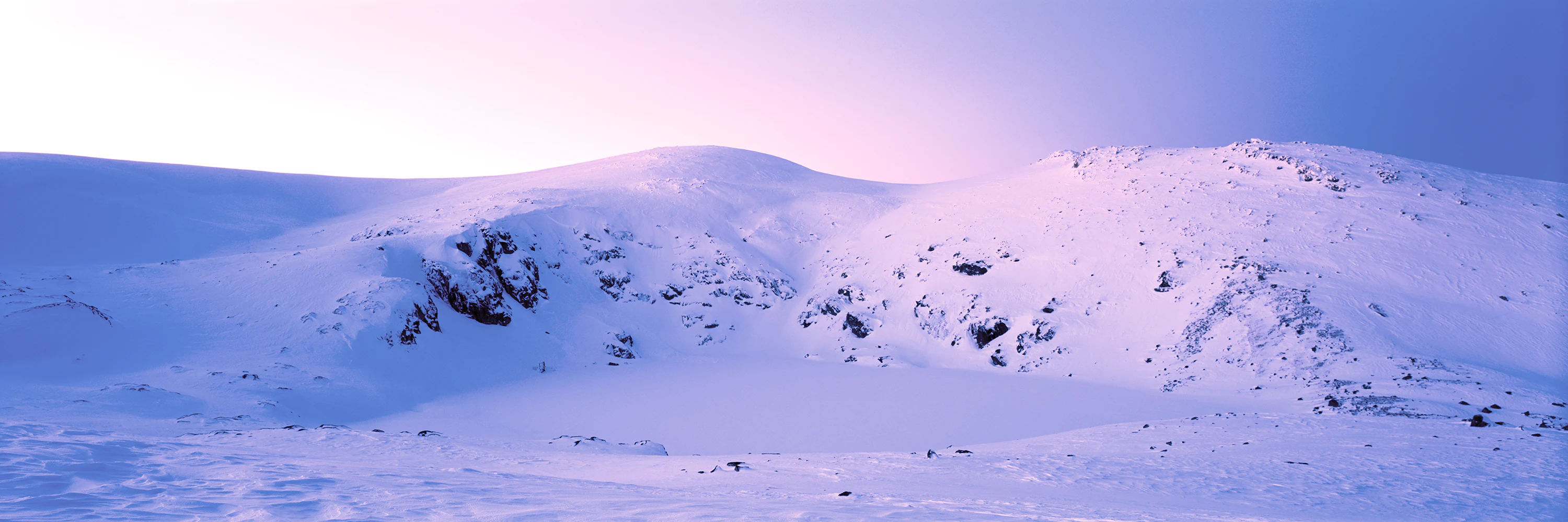 Cordillera montañosa nevada con afloramientos rocosos bajo un cielo rosa pálido