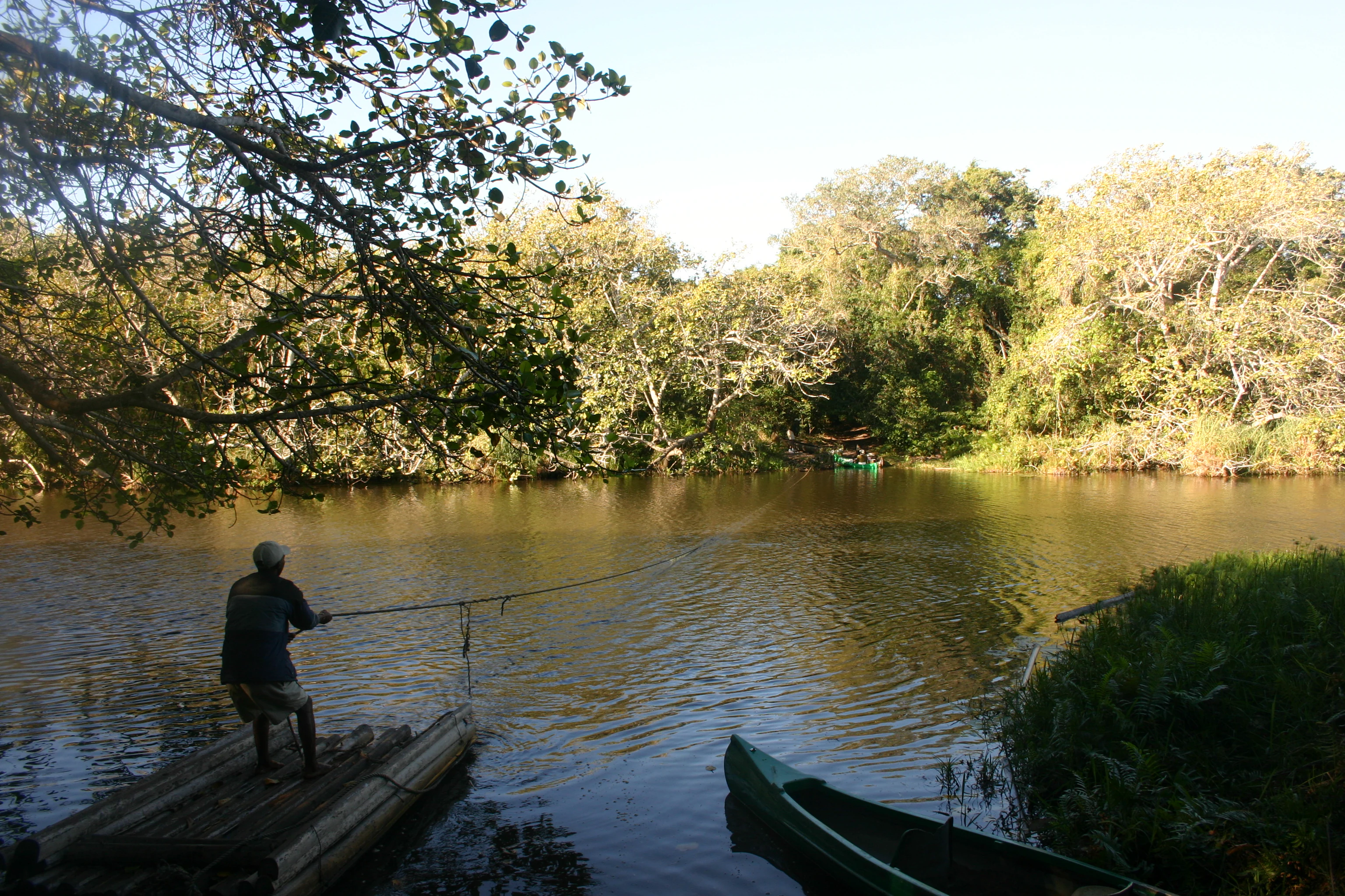 Una persona está de pie en una pequeña balsa de madera en un río o lago tranquilo, tirando de una línea de pesca, con una canoa cerca y árboles densos bordeando la orilla.