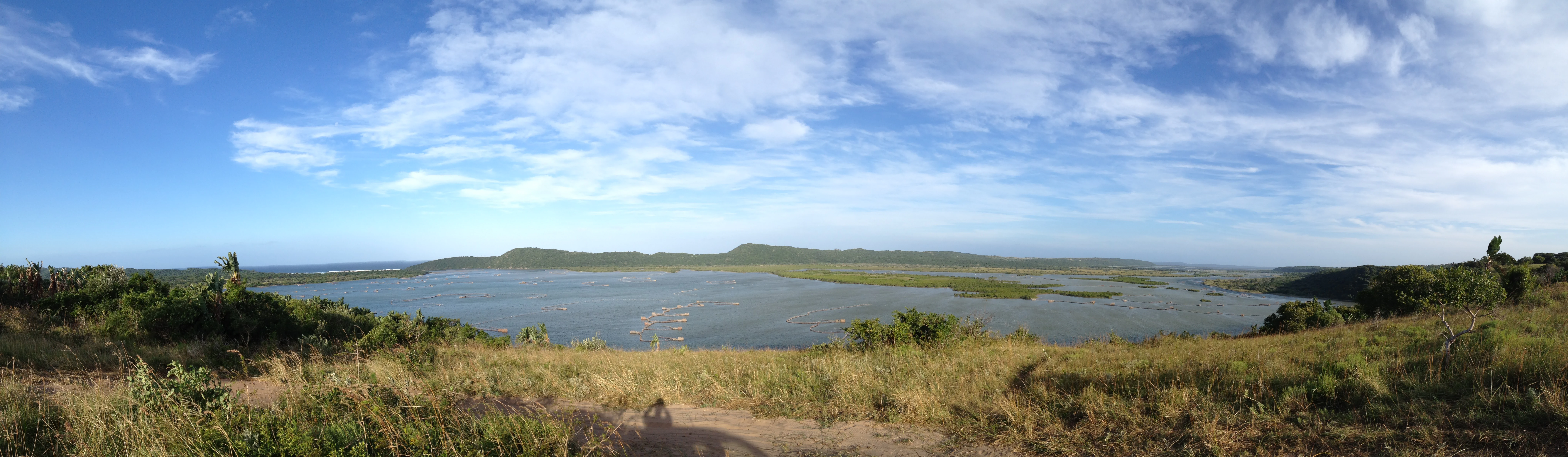 Una amplia toma panorámica del estuario de la bahía de Kosi, con una extensión tranquila de agua que se extiende hacia colinas verdes lejanas bajo un cielo azul brillante y nublado.