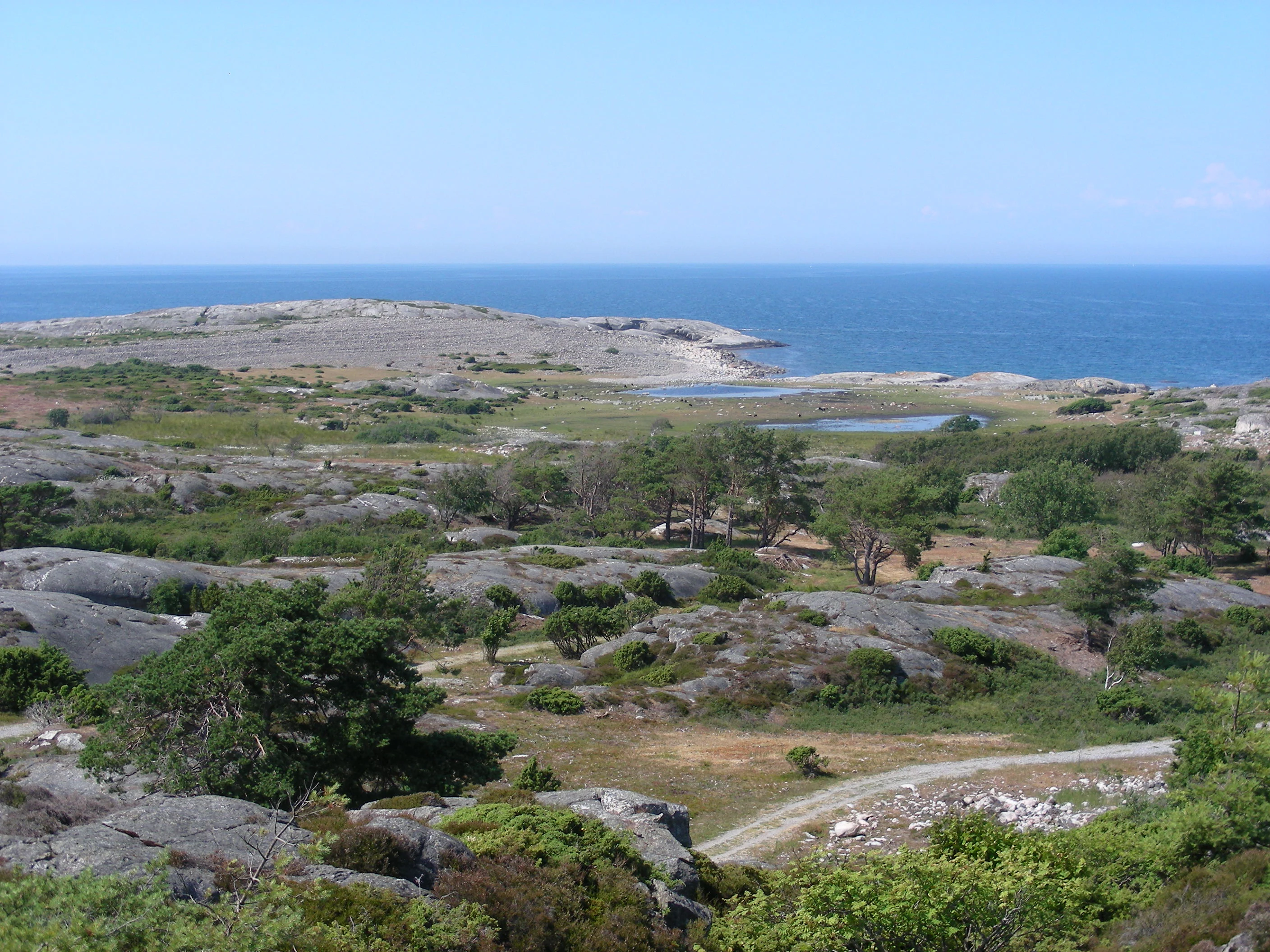 Rocky terrain with scattered vegetation, a winding path, and the sea visible under a clear blue sky
