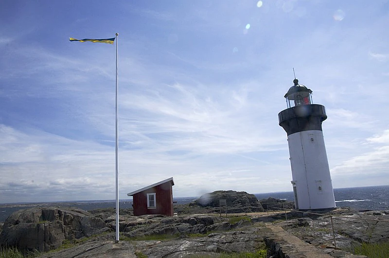 White lighthouse with black top, red building, tall flagpole with yellow flag, rocky terrain, and ocean in background under blue sky with clouds