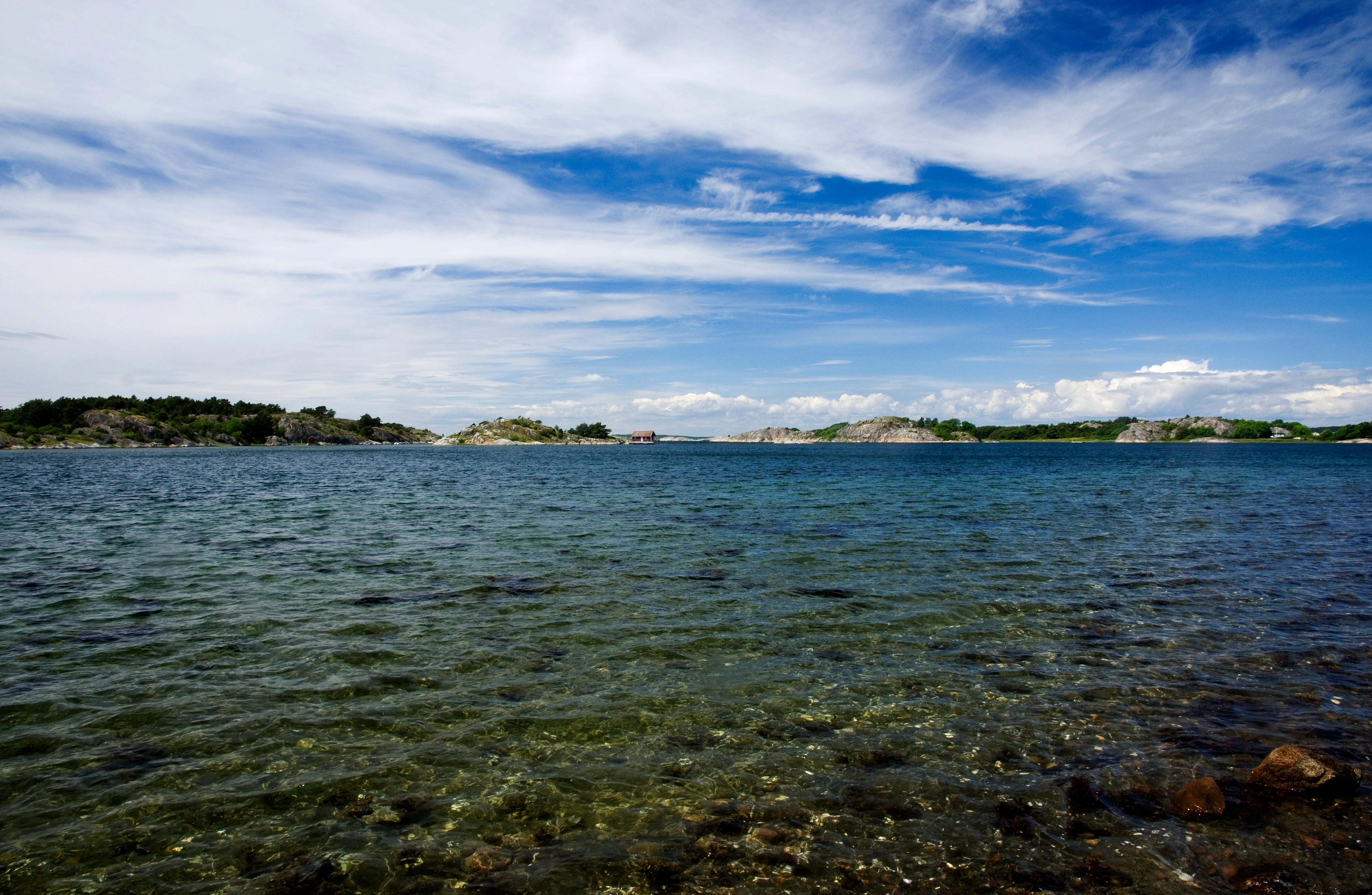 Wide view of calm blue water with rocky shoreline in foreground and small islands visible in the distance under a partly cloudy sky