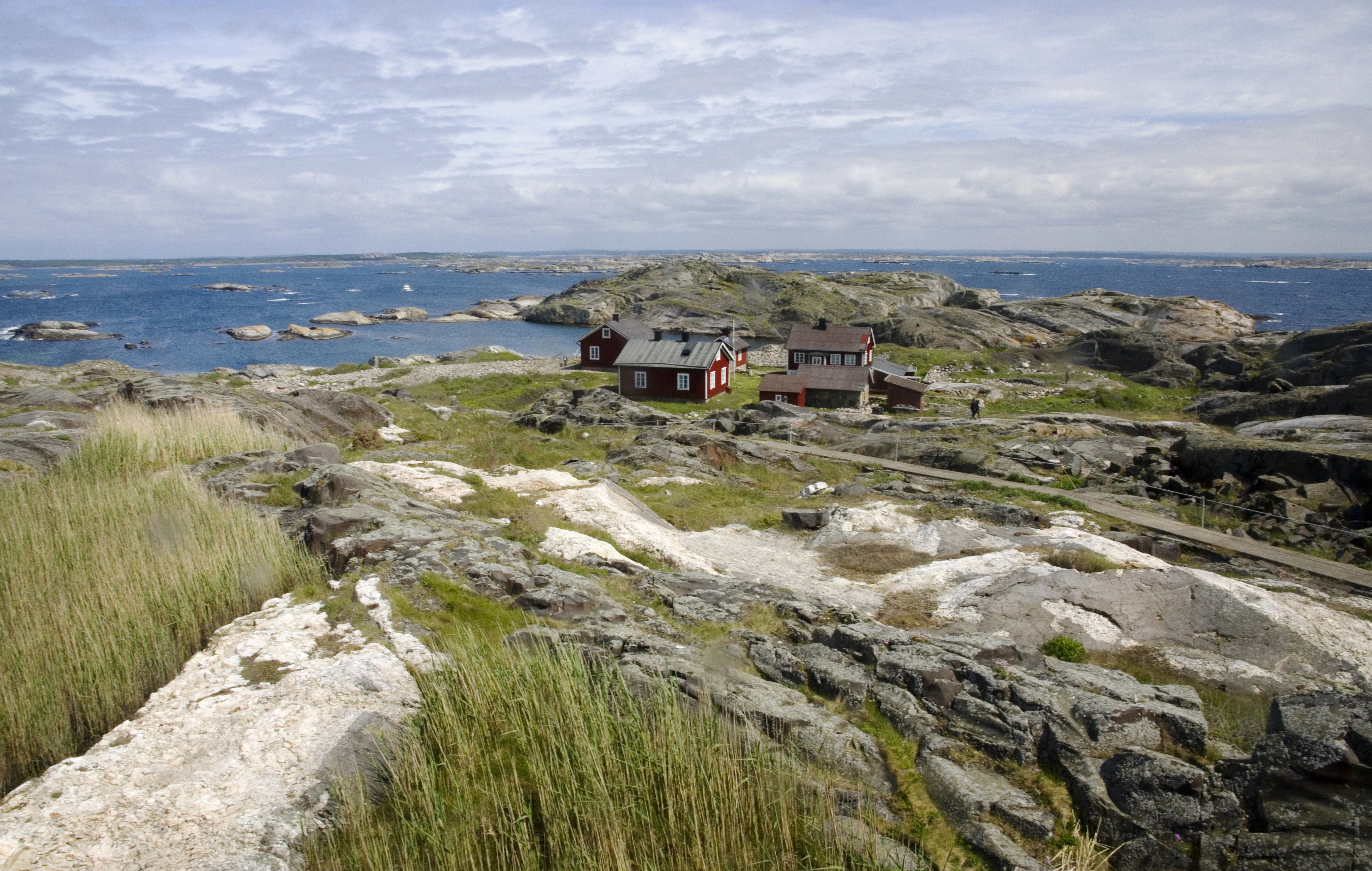 Rocky coastal terrain with grassy areas, red houses, and distant islands in the sea under a partly cloudy sky.
