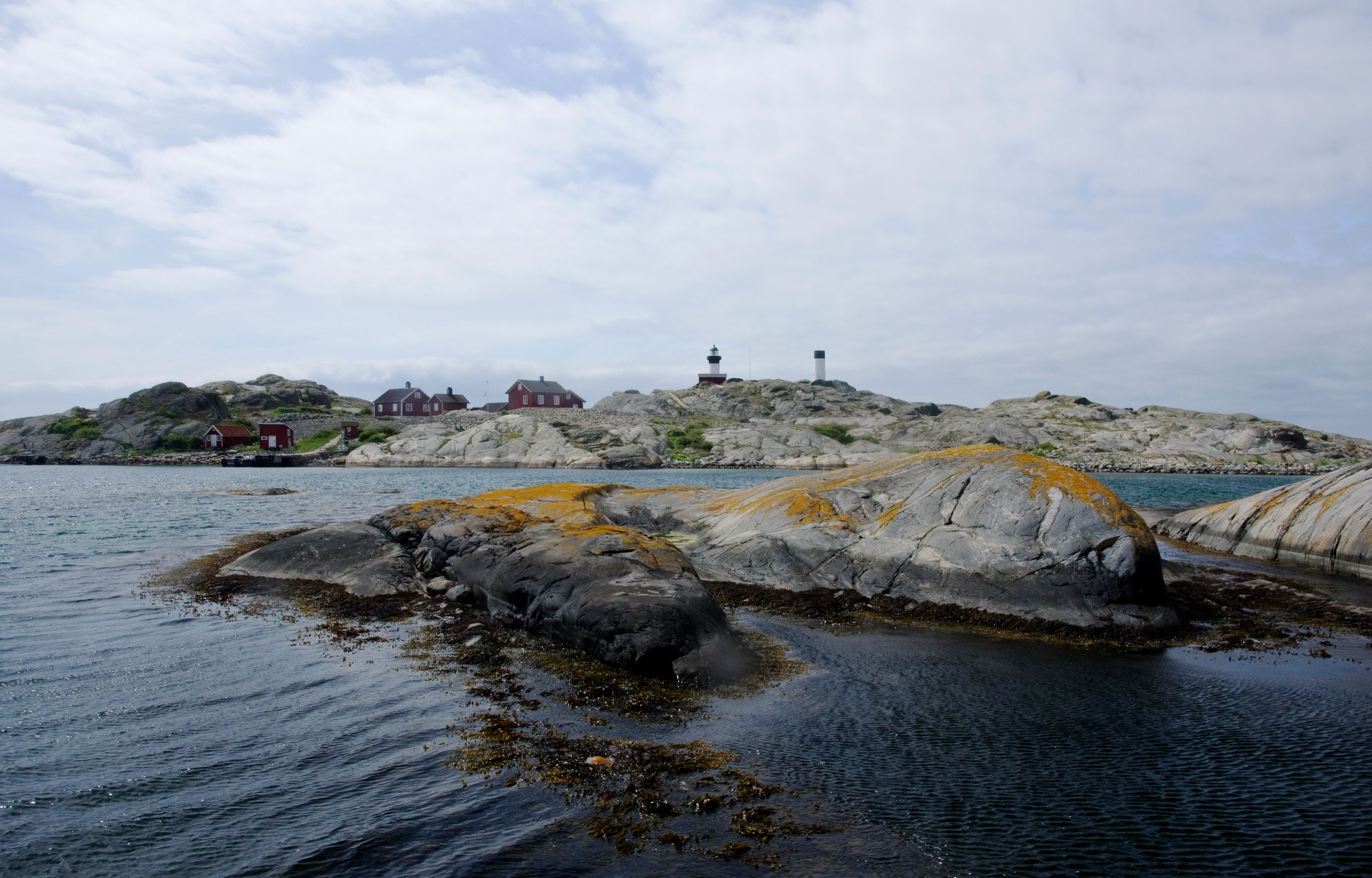 Rocky islands covered in lichen with calm water surrounding them and distant buildings on a larger island under a cloudy sky