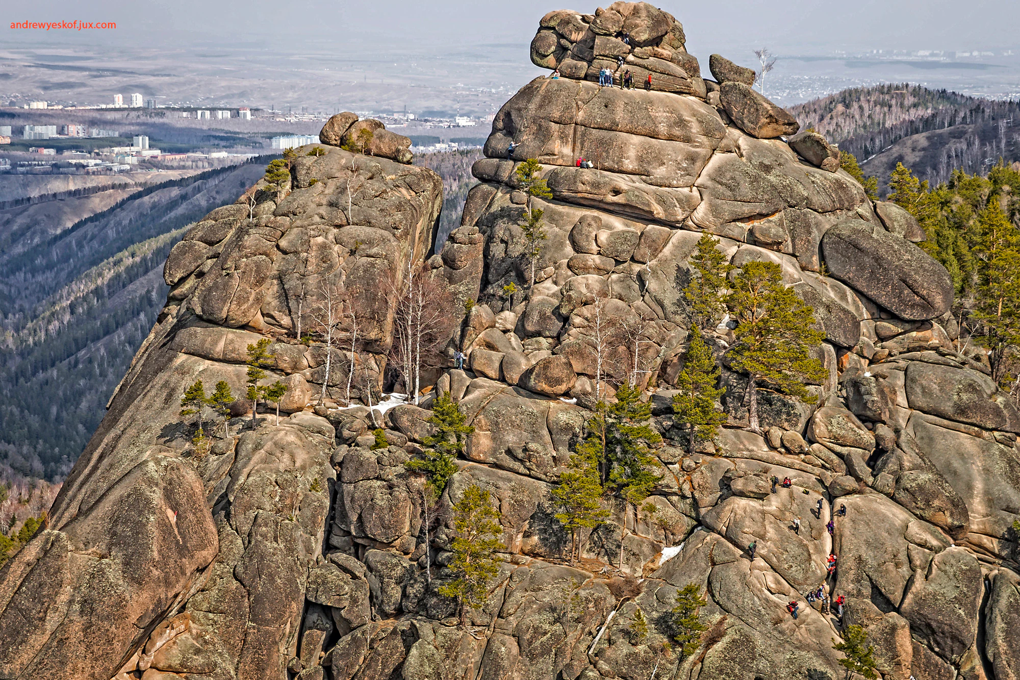 Large rocky mountain formations with scattered coniferous trees, small figures of people climbing, distant valley, and urban area visible in background