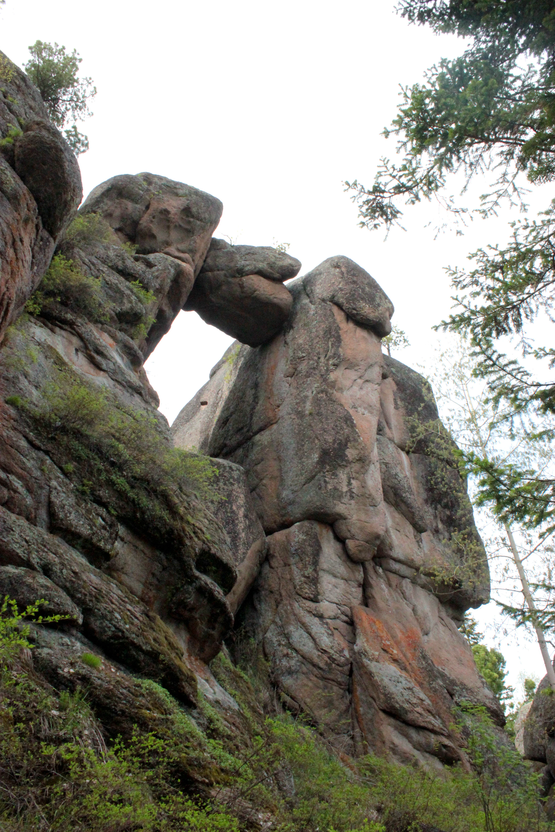 A low angle view of a large natural rock pillar formation with a smaller stone wedged between two larger columns, creating a gate-like arch, set against a bright sky with pine branches.