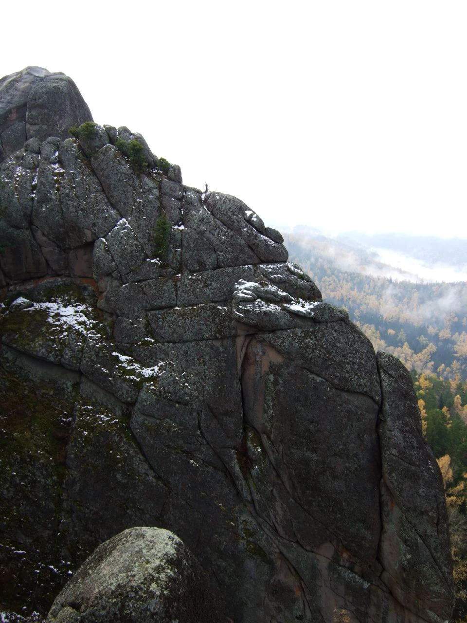 A close-up view of a large, weathered, grey syenite rock pillar with small patches of snow, overlooking a forested valley partially obscured by mist.