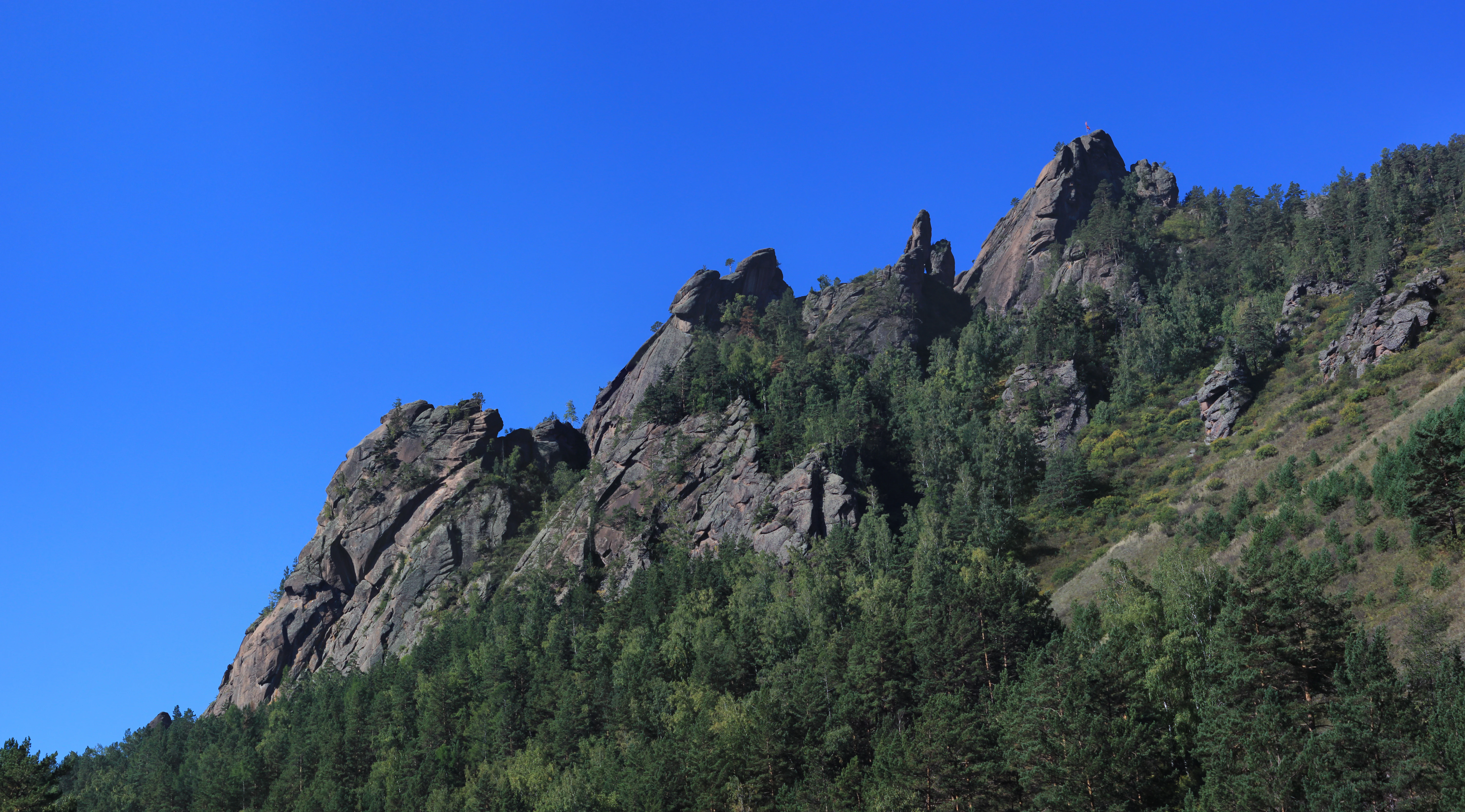 A jagged, rocky mountain formation known as Takmak rising above a dense green pine forest under a bright clear blue sky.