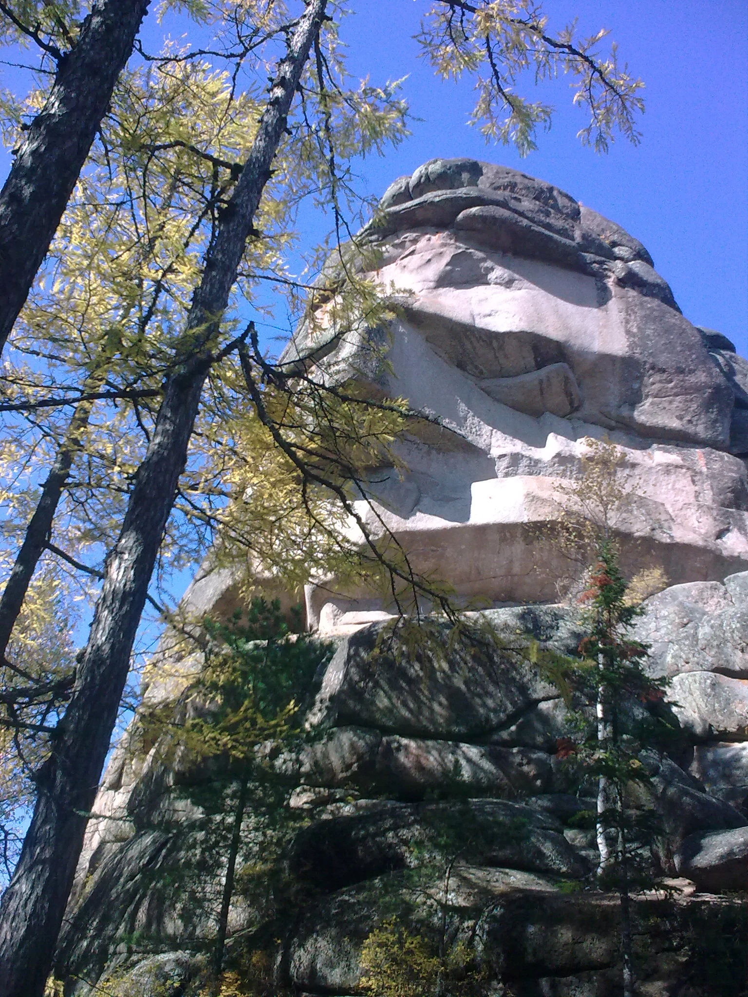A low angle view of the massive Ded syenite rock pillar rising above coniferous trees under a clear blue sky.