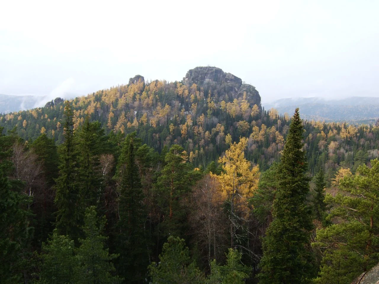 A wide view of a forested hillside with golden and dark green trees, featuring a prominent, craggy rock pillar at the summit under an overcast sky.