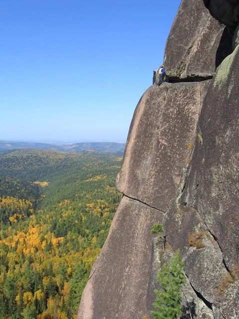 A close up view of a sheer, weathered rock pillar with a person climbing near the top, overlooking a vast, dense forest canopy showing autumn foliage colors.