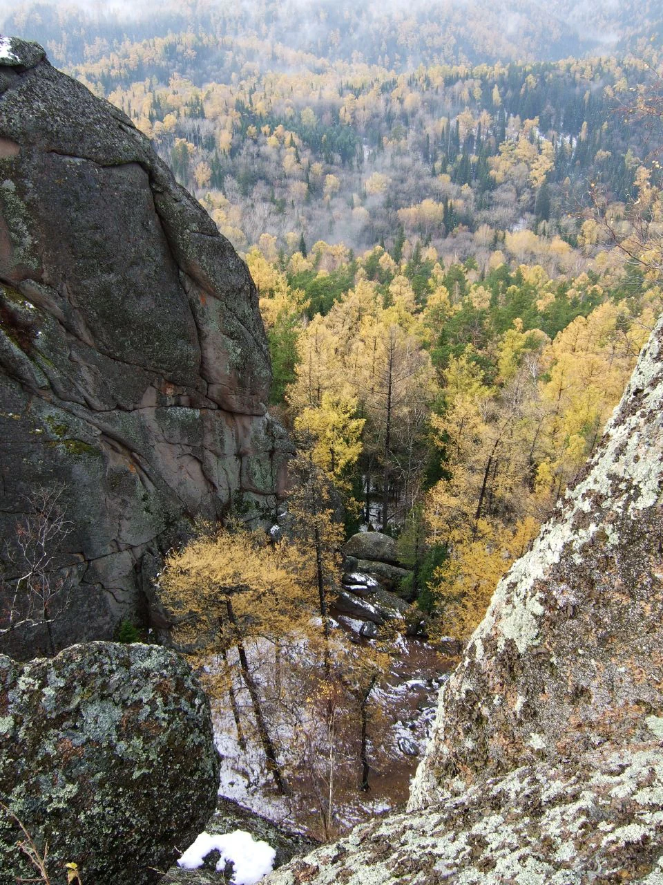 A high-angle view looking down between two large, weathered grey rock pillars into a dense forest valley with yellow autumn trees and patches of early snow.